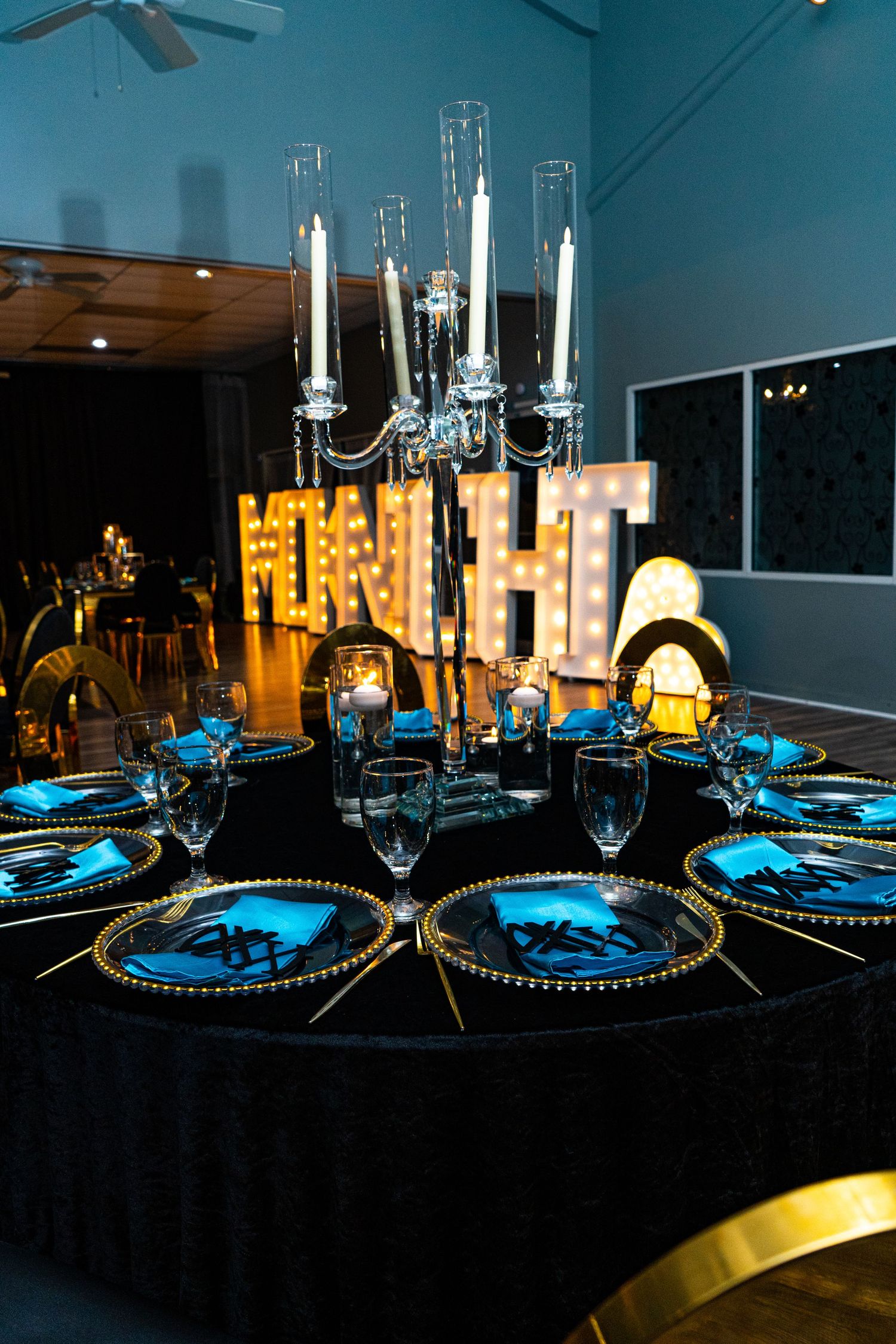 Elegant party table setting with black tablecloth, crystal stemware, illuminated marquee letters and glass candelabra centerpiece.