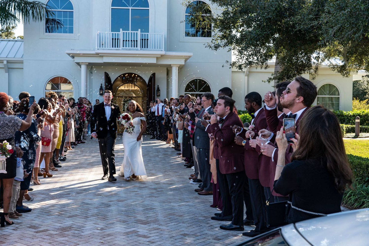 Wedding guests line a brick walkway outside a white mansion as newlyweds exit through elegant arched doorways.
