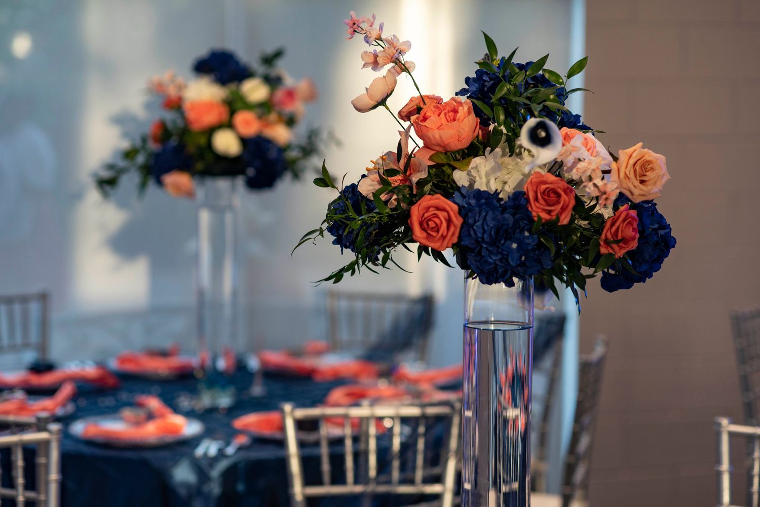 Floral centerpiece with coral roses and navy blue hydrangeas in tall glass vase at elegant wedding reception table setting.