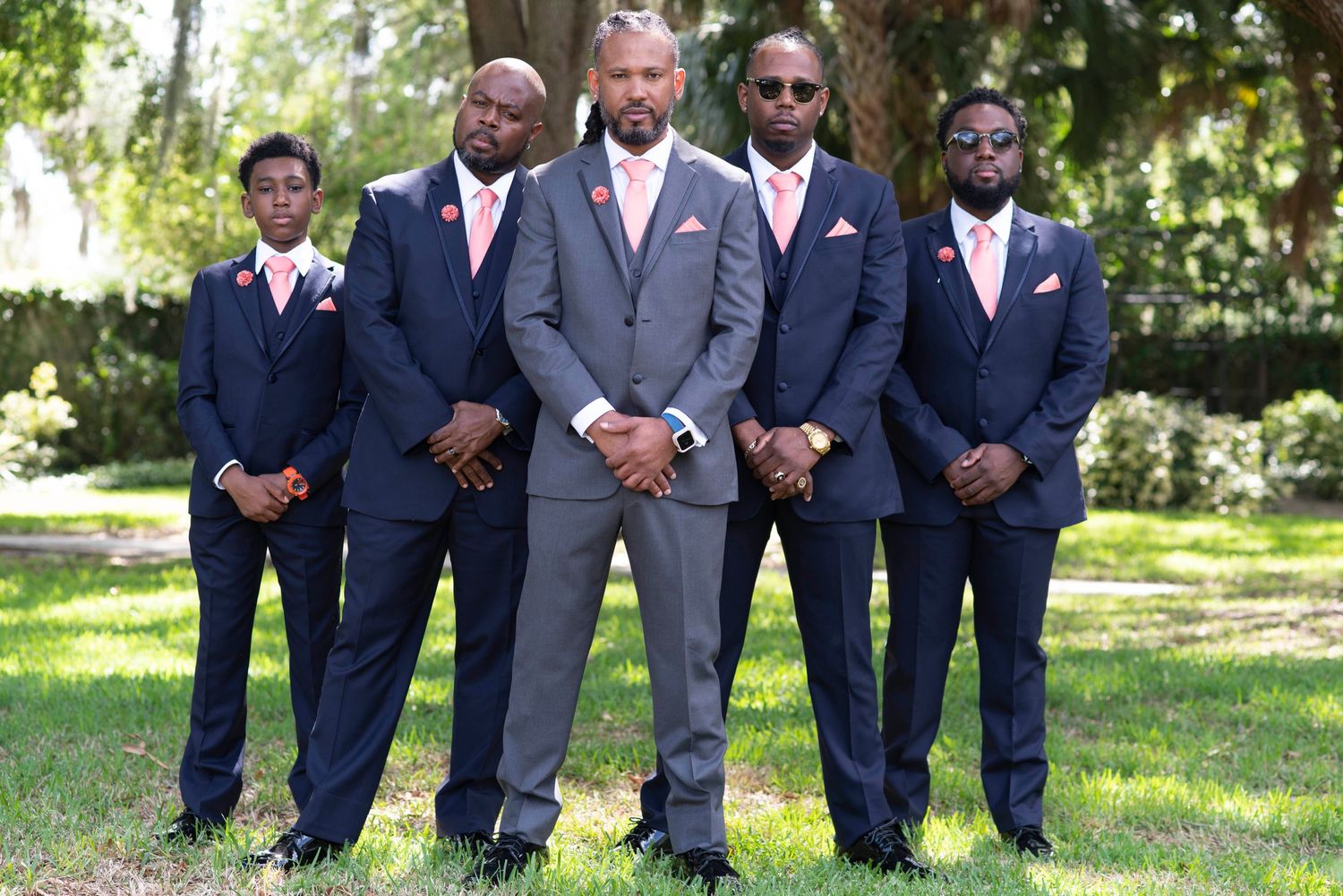 A group of groomsmen pose together outdoors wearing coordinated navy blue and grey suits with pink accents.