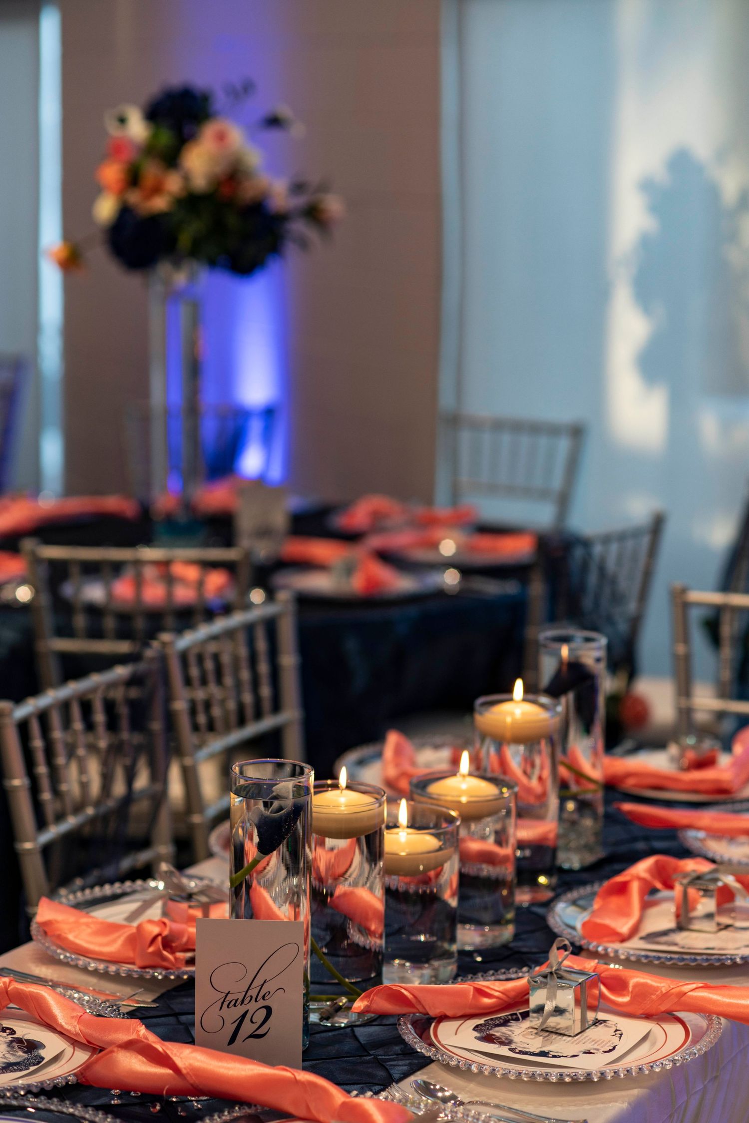 Elegant wedding reception table set with navy linens, coral napkins, and glowing candle centerpieces in a dimly lit room.