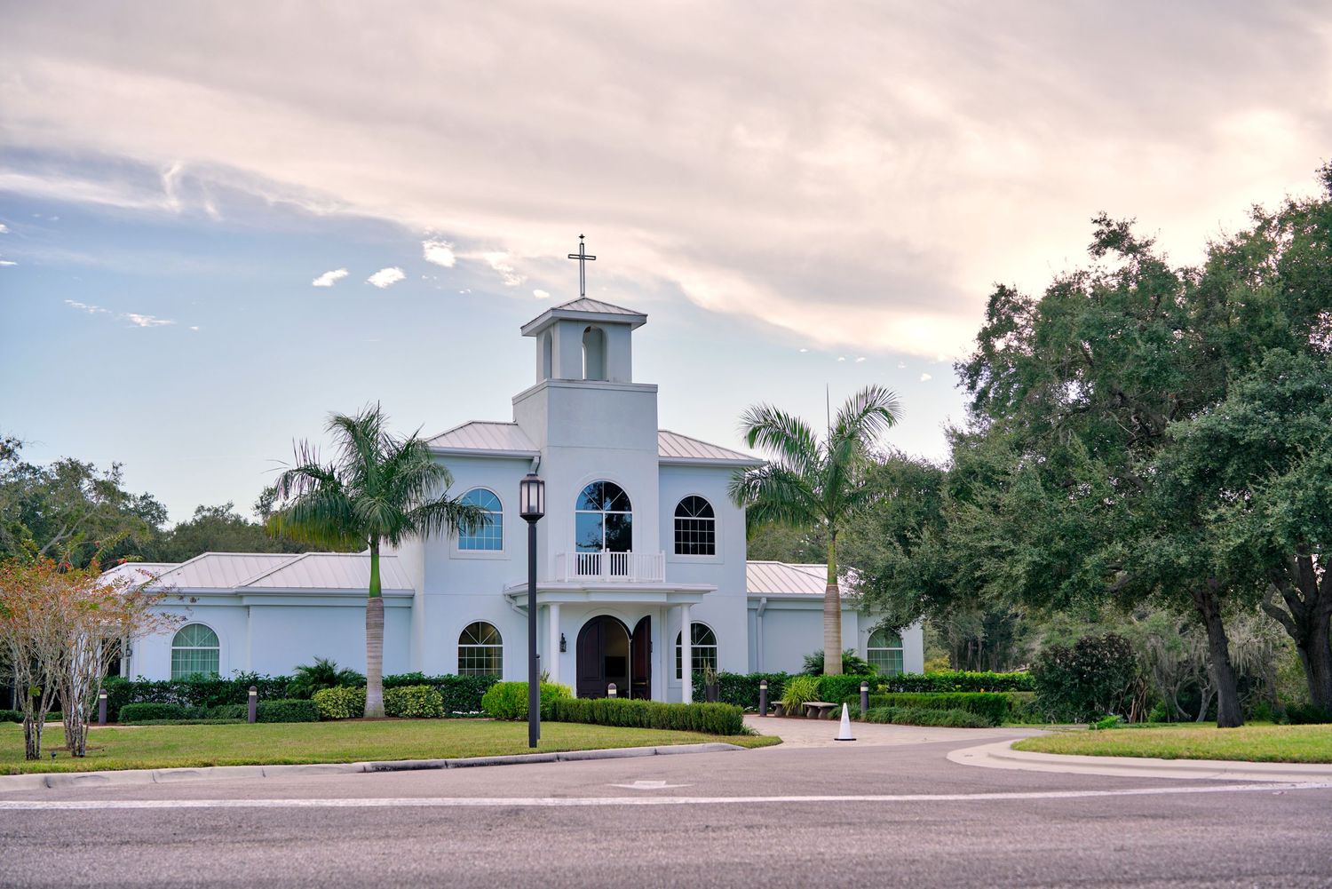 White Spanish-style church building with bell tower and palm trees against cloudy sky in Florida.