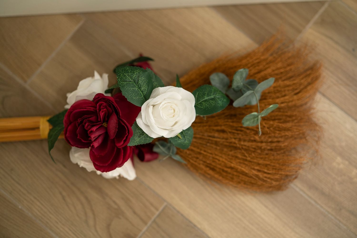 A decorative broom made with natural straw and adorned with red and white roses and green leaves on a wooden floor.