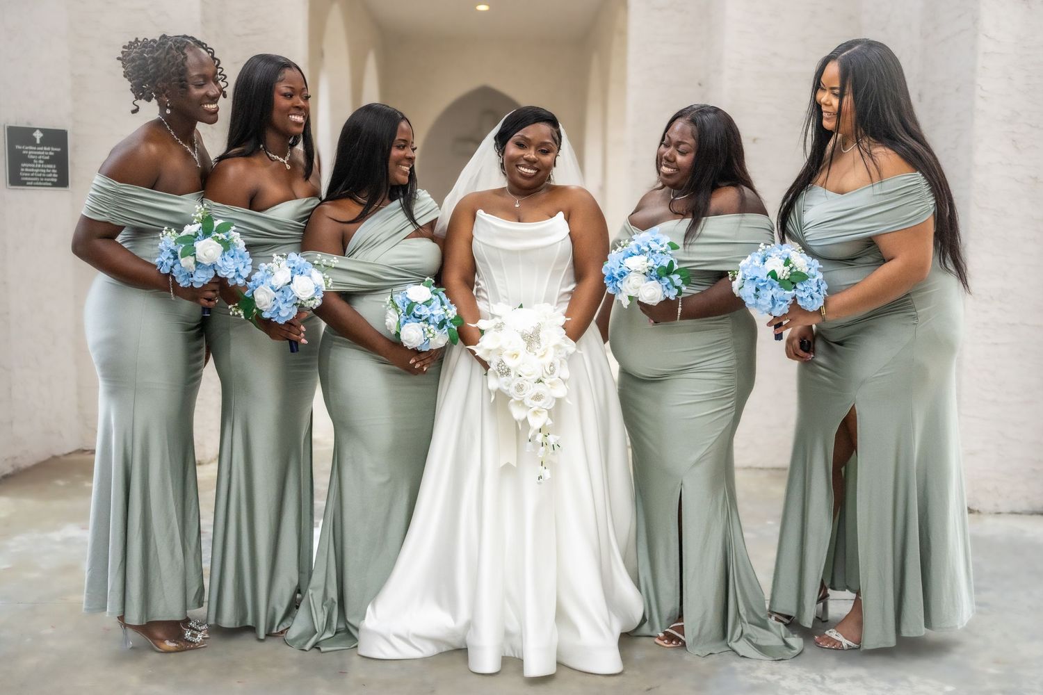 Bridesmaids in sage green dresses pose with the bride in a white wedding gown.