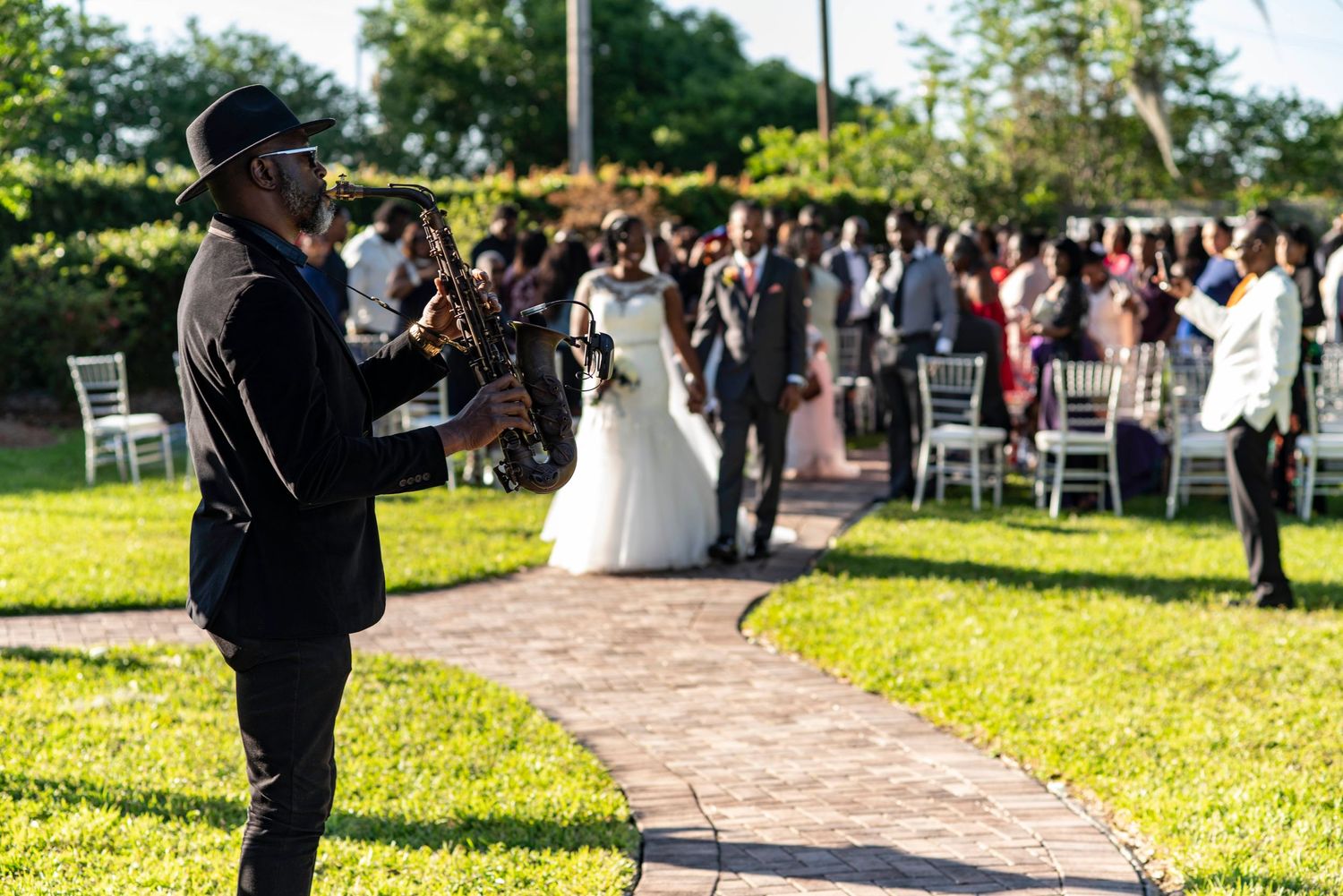 Wedding photographer takes photos during an outdoor ceremony on a stone path surrounded by green lawn and guests.
