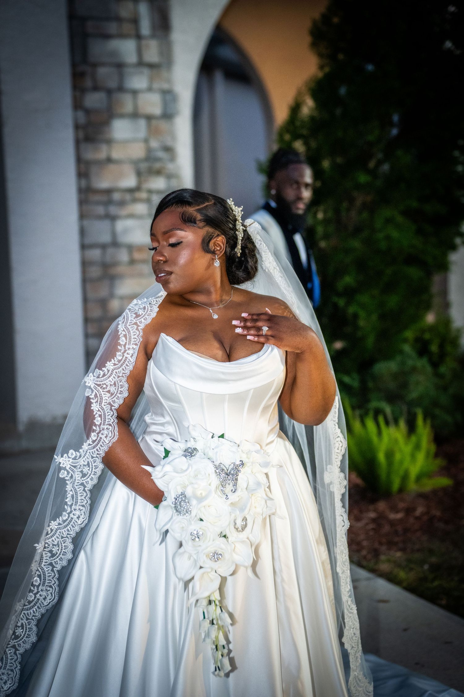 Beautiful wedding photo with a white satin gown, lace veil and cascading orchid bouquet outside a stone archway.