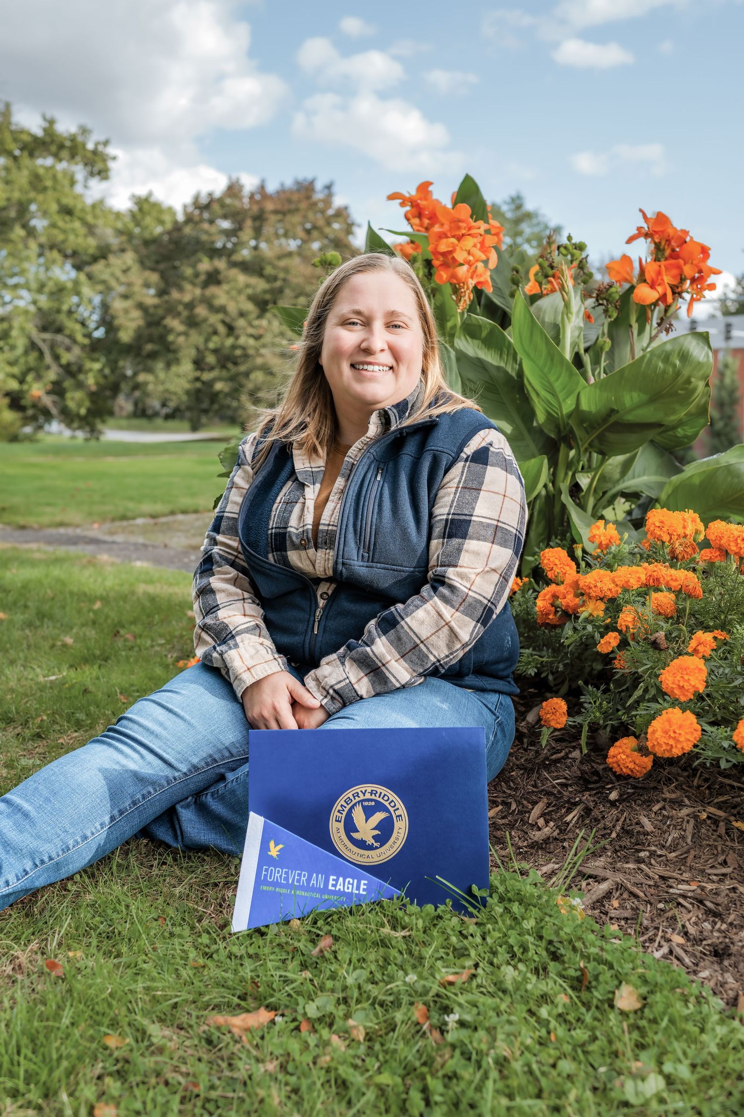 Someone sits on grass next to vibrant orange flowers and a blue box on a sunny day wearing plaid and a vest.