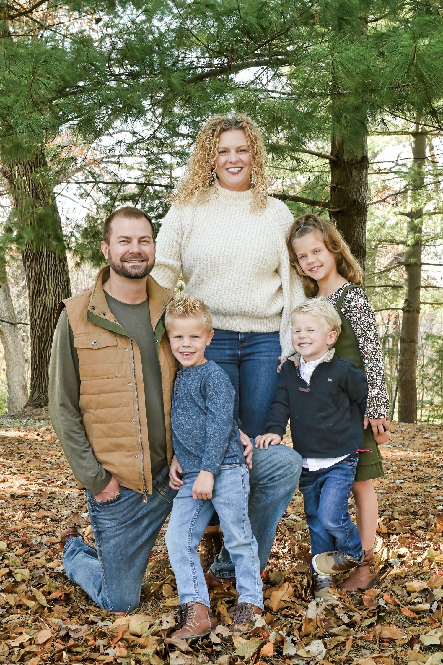 A family of five pose together outdoors on fallen autumn leaves among tall evergreen trees in a wooded setting.