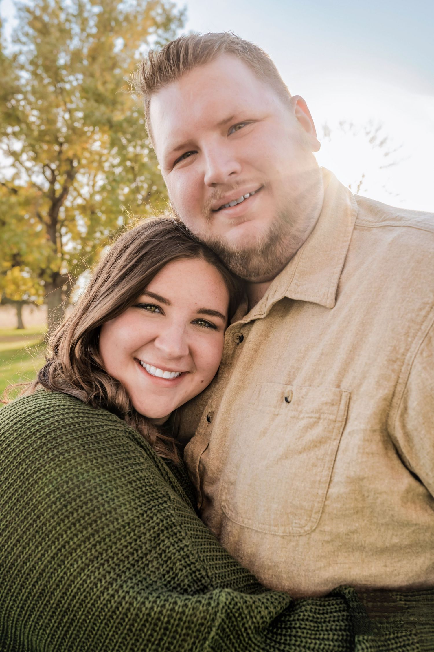 A couple embraces in an outdoor fall setting with autumn trees and sunlight in the background.