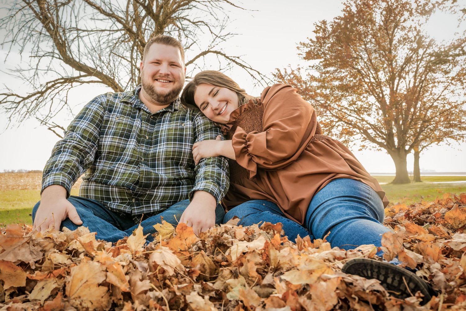 A couple sits together outdoors in a pile of autumn leaves surrounded by bare trees on a sunny fall day.