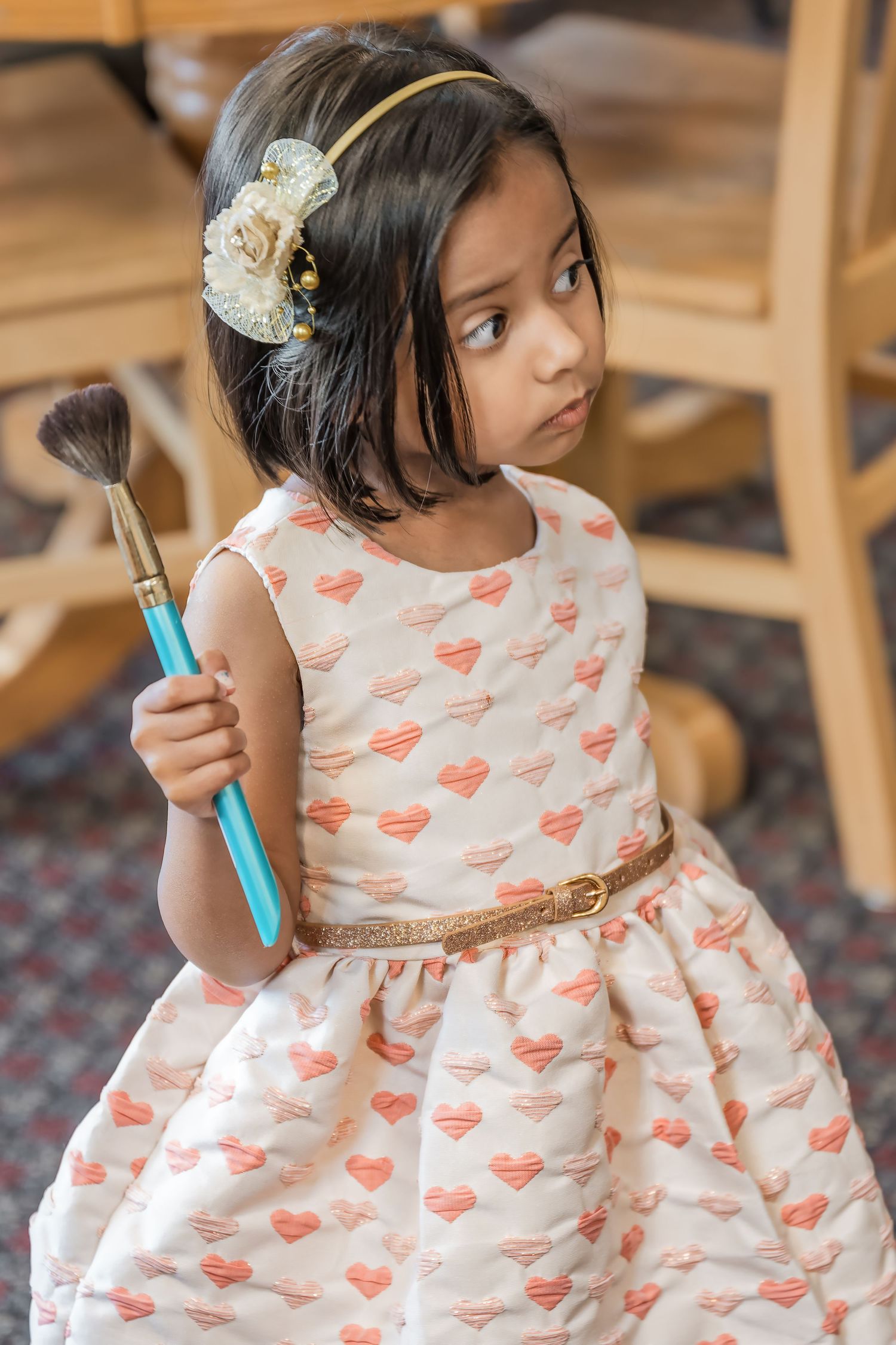 Girl in heart-patterned dress holds makeup brush while wearing flower hair accessory.