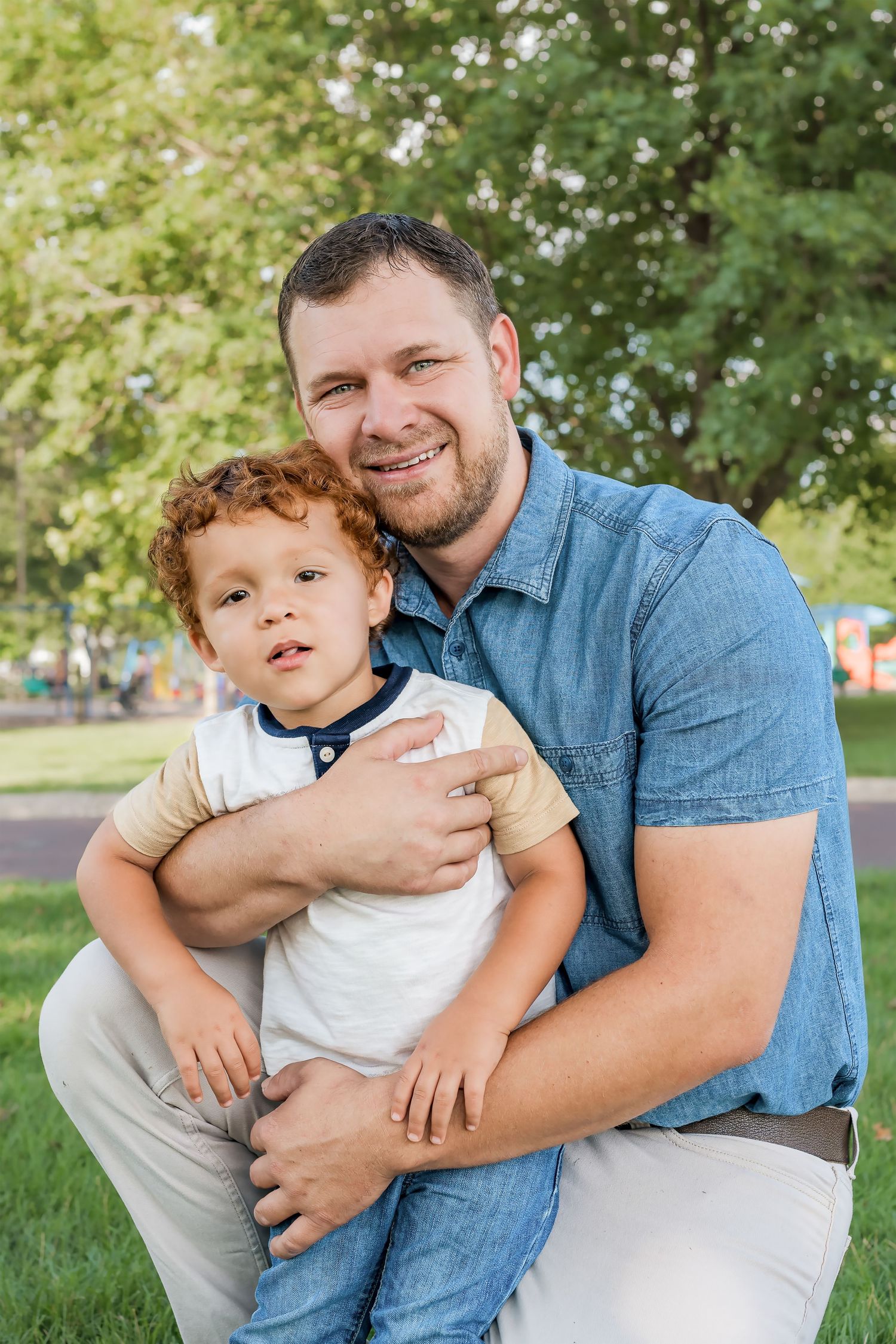 A heartwarming outdoor portrait of a parent and child with curly red hair embracing on a sunny day in a park setting.