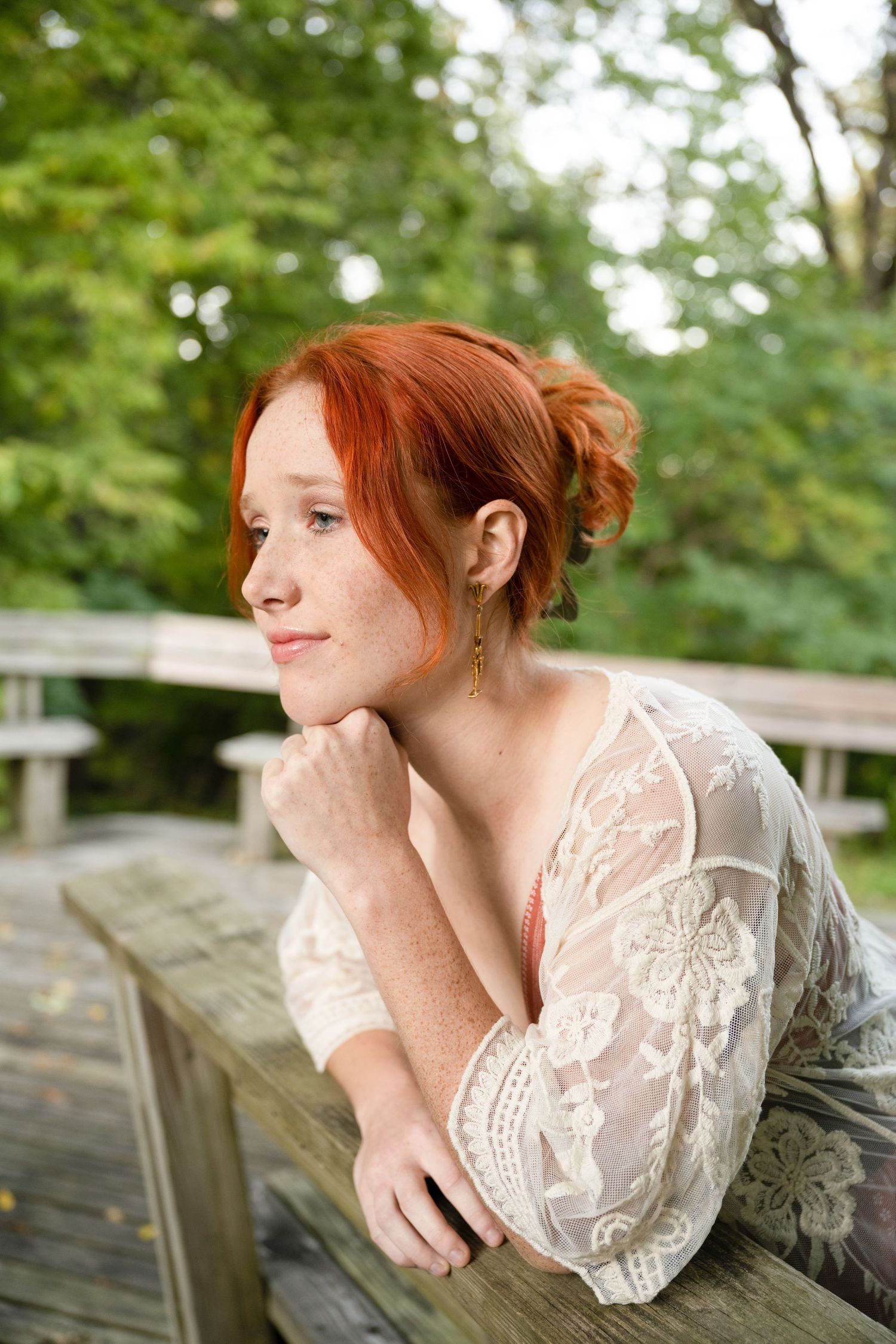 A serene portrait in a lace dress leaning thoughtfully on a wooden deck railing with green foliage in the background.