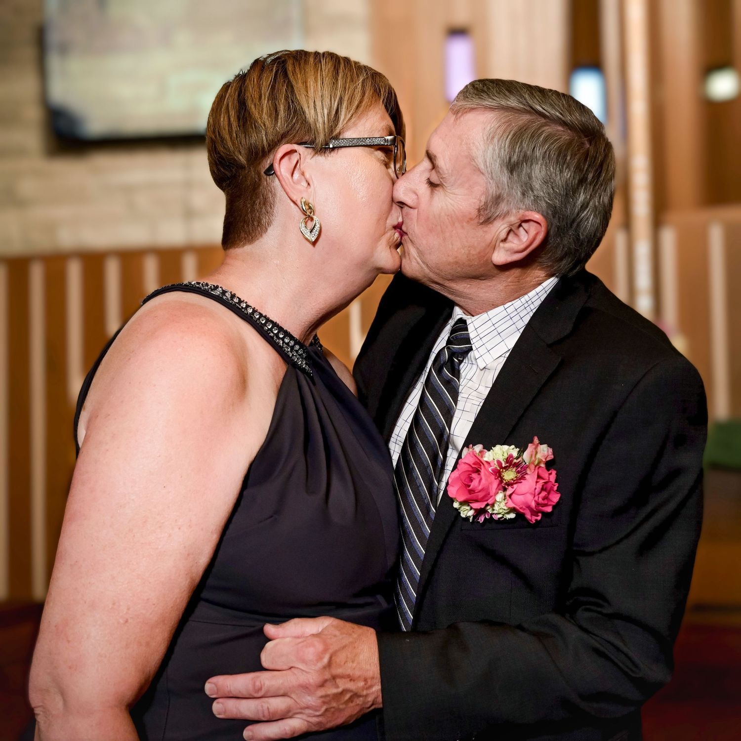 A tender kiss shared between a couple at a formal event, with a pink floral boutonniere adorning the suit.