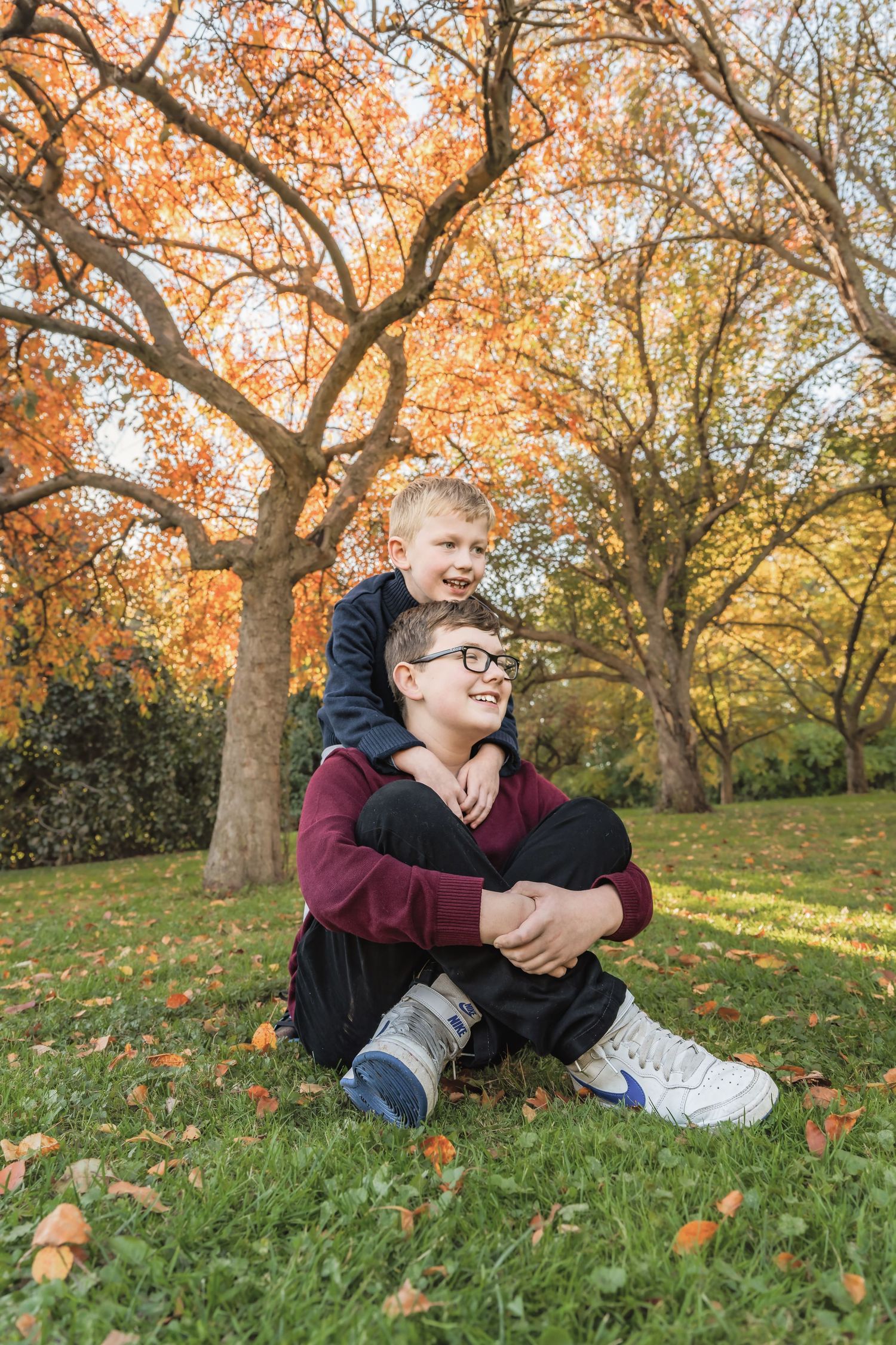 Two people sit together on autumn grass surrounded by fall foliage and trees with warm orange and yellow leaves.