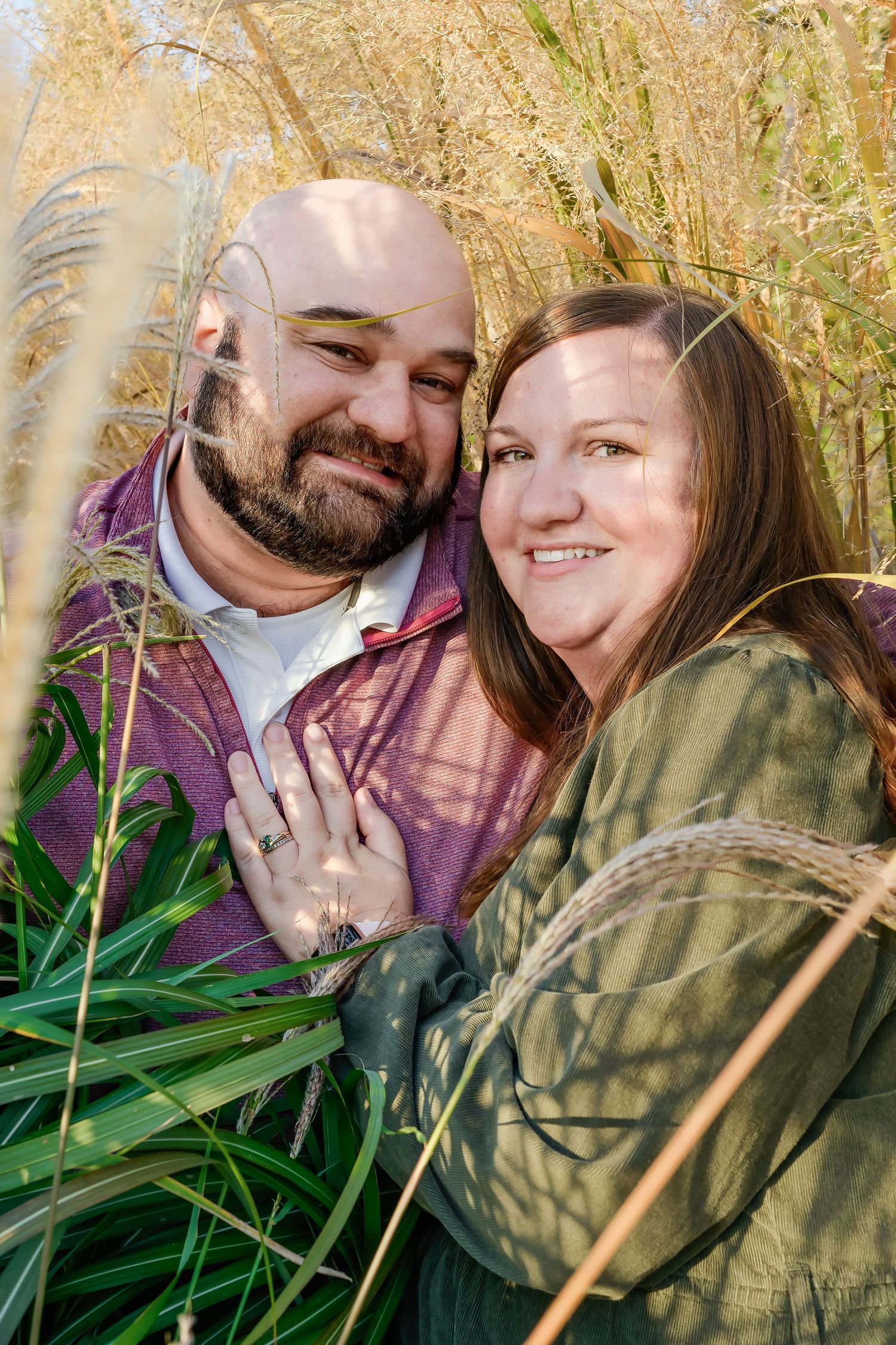 A couple smiles together outdoors among tall golden grasses while wearing casual outerwear.