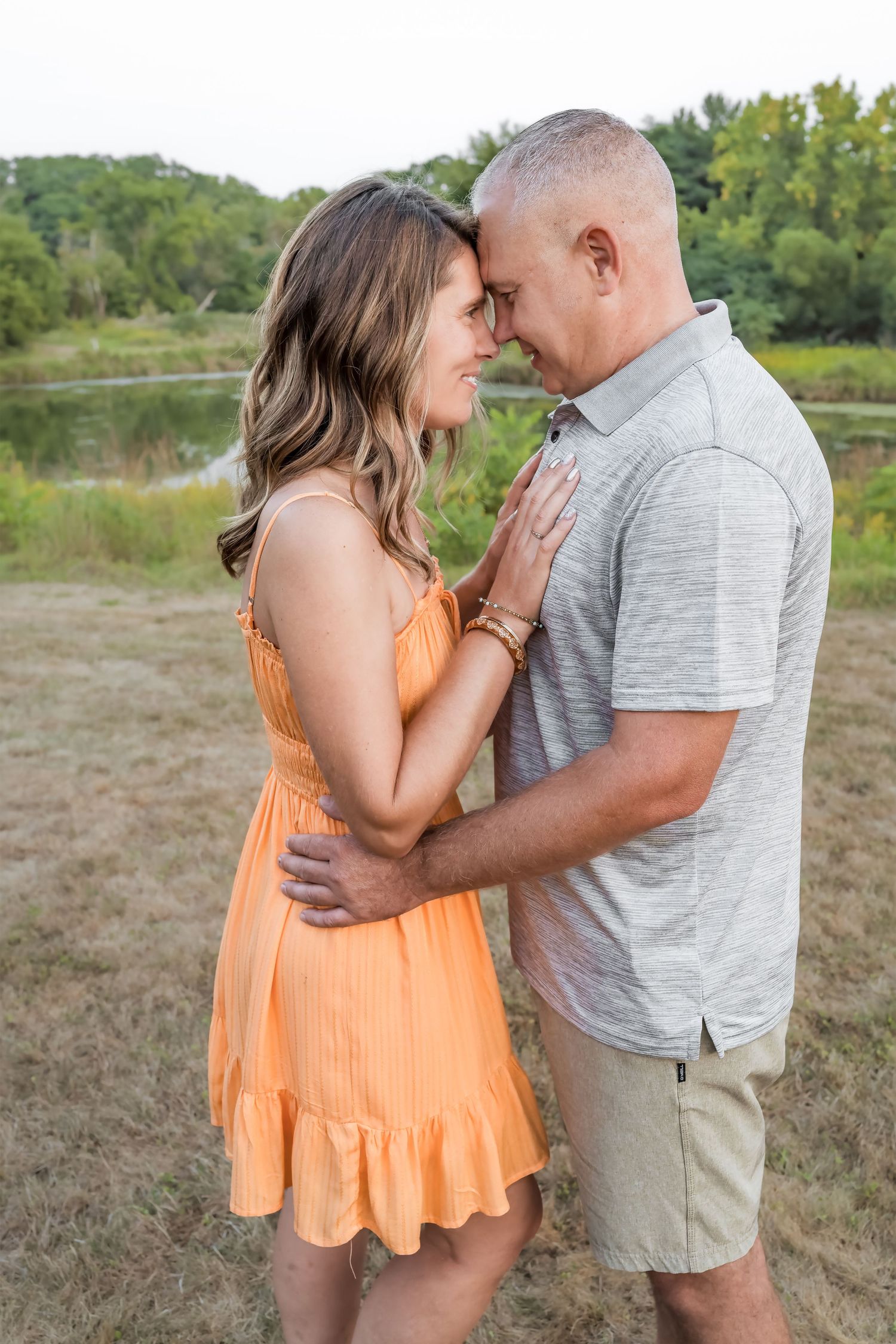 A couple embraces intimately outdoors near a lake, with her wearing a peach-colored dress and him in a gray polo shirt.