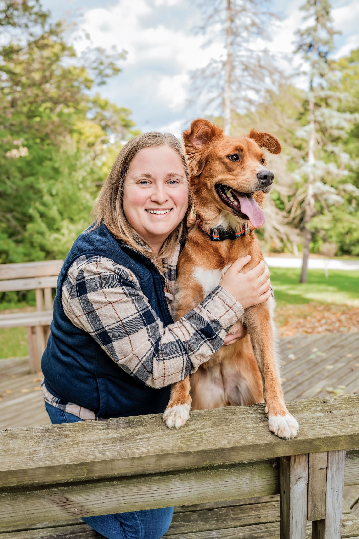Happy dog with reddish-brown fur stands on wooden bridge railing with companion in plaid and navy vest outdoors.