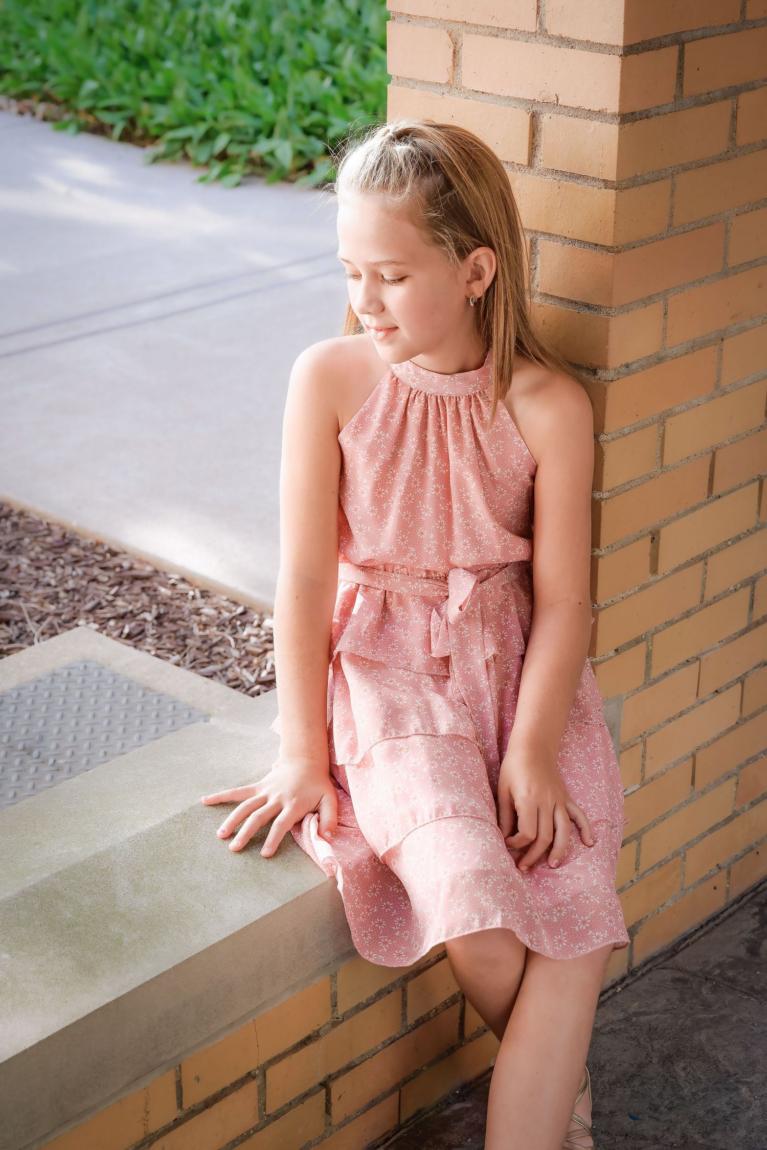 Girl in pink summer dress sits thoughtfully on brick ledge outdoors.