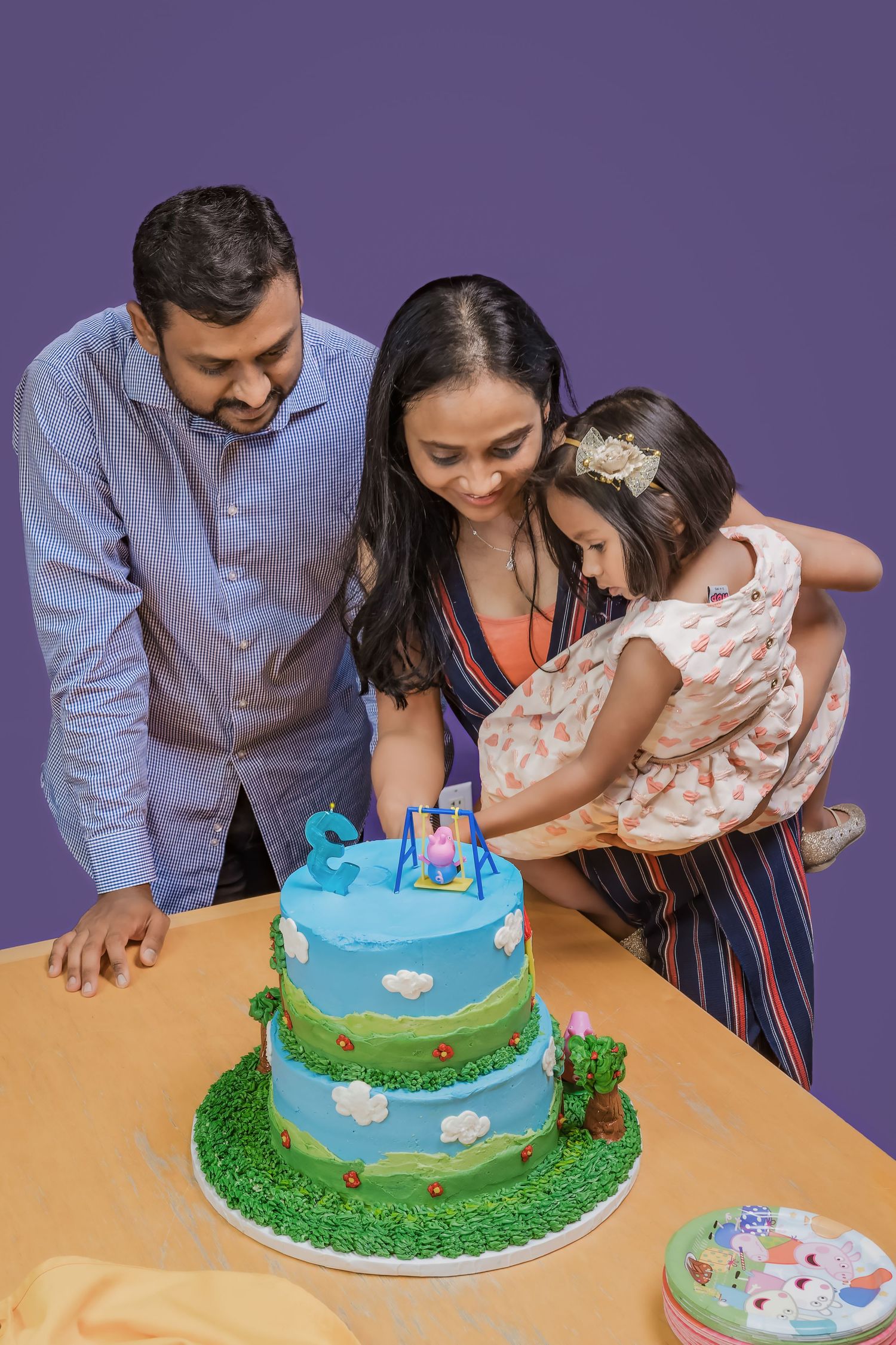 A family gathers around a blue two-tiered birthday cake decorated with clouds and grass details on an orange tablecloth.
