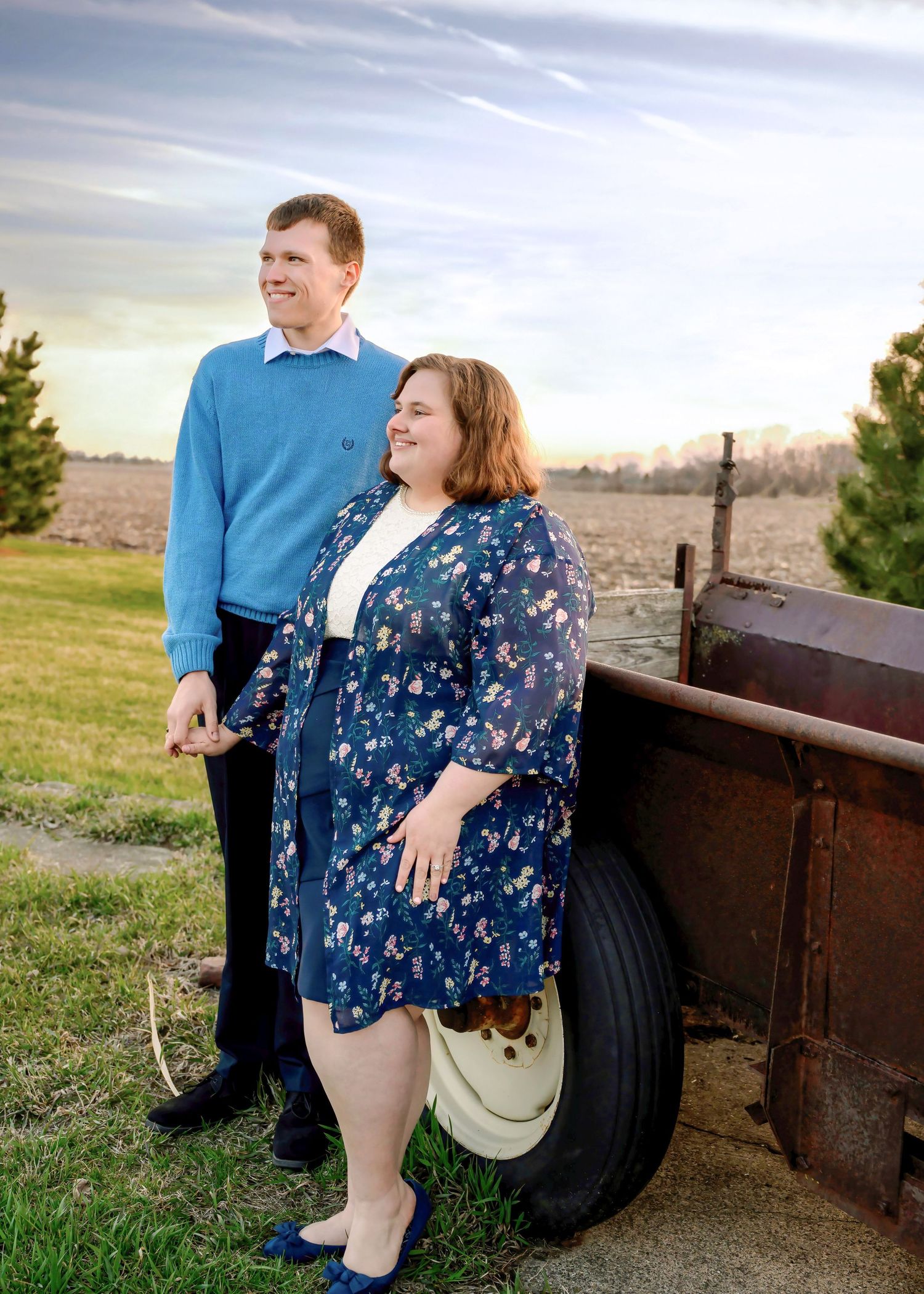Two people smile while posing together in a rural setting with an old rustic farm equipment wheel visible.