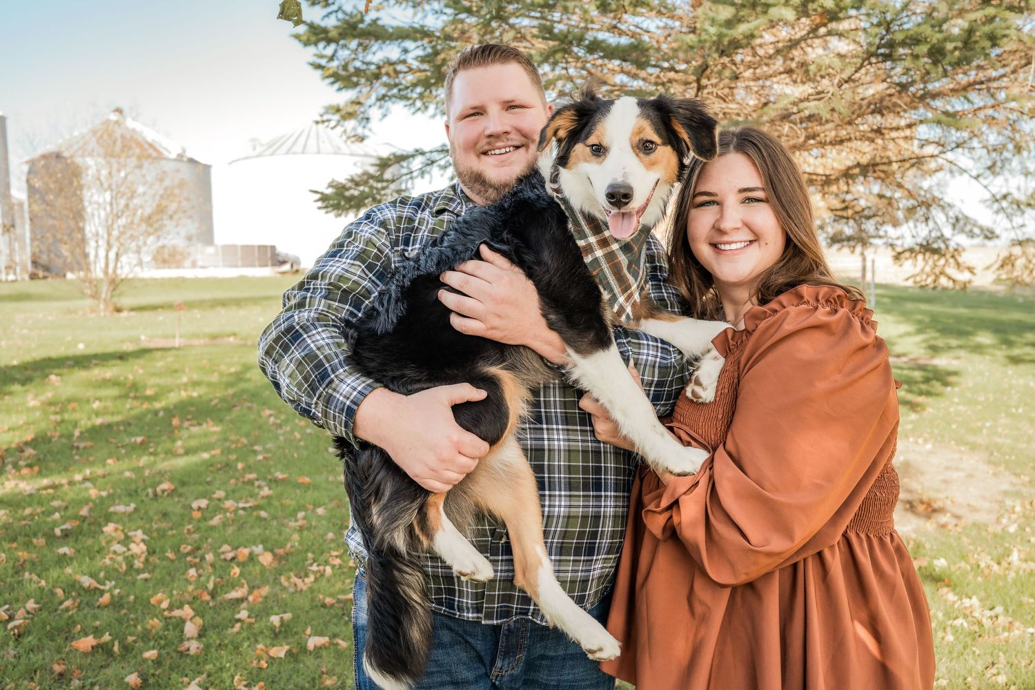 Happy couple outdoors holding their smiling Australian Shepherd dog on a sunny autumn day.