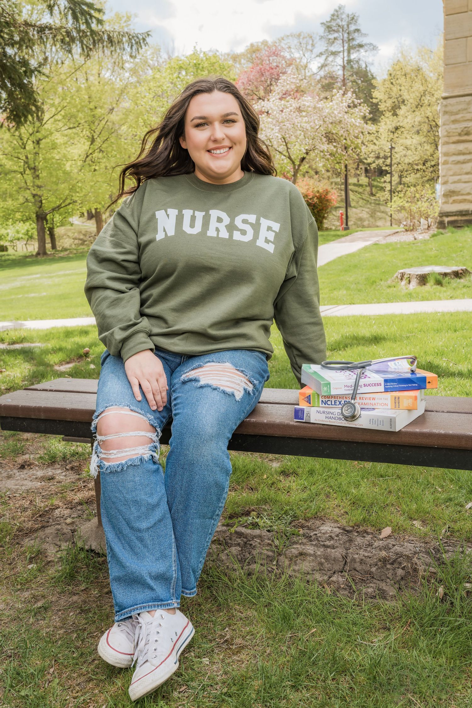 Healthcare student sitting on bench wearing green nurse sweatshirt with textbooks and medical supplies outdoors.