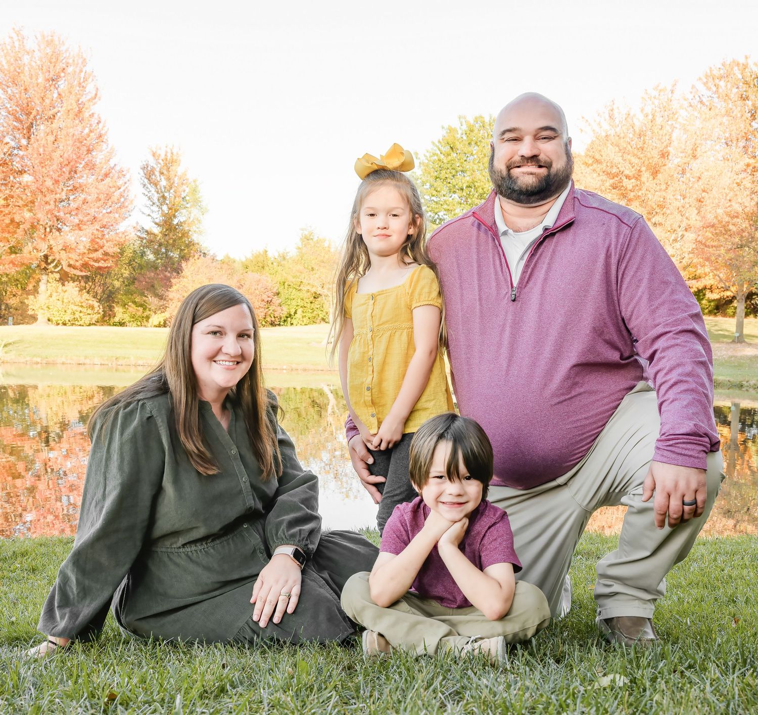 A family poses together outdoors on grass with autumn trees showing fall colors in the background.