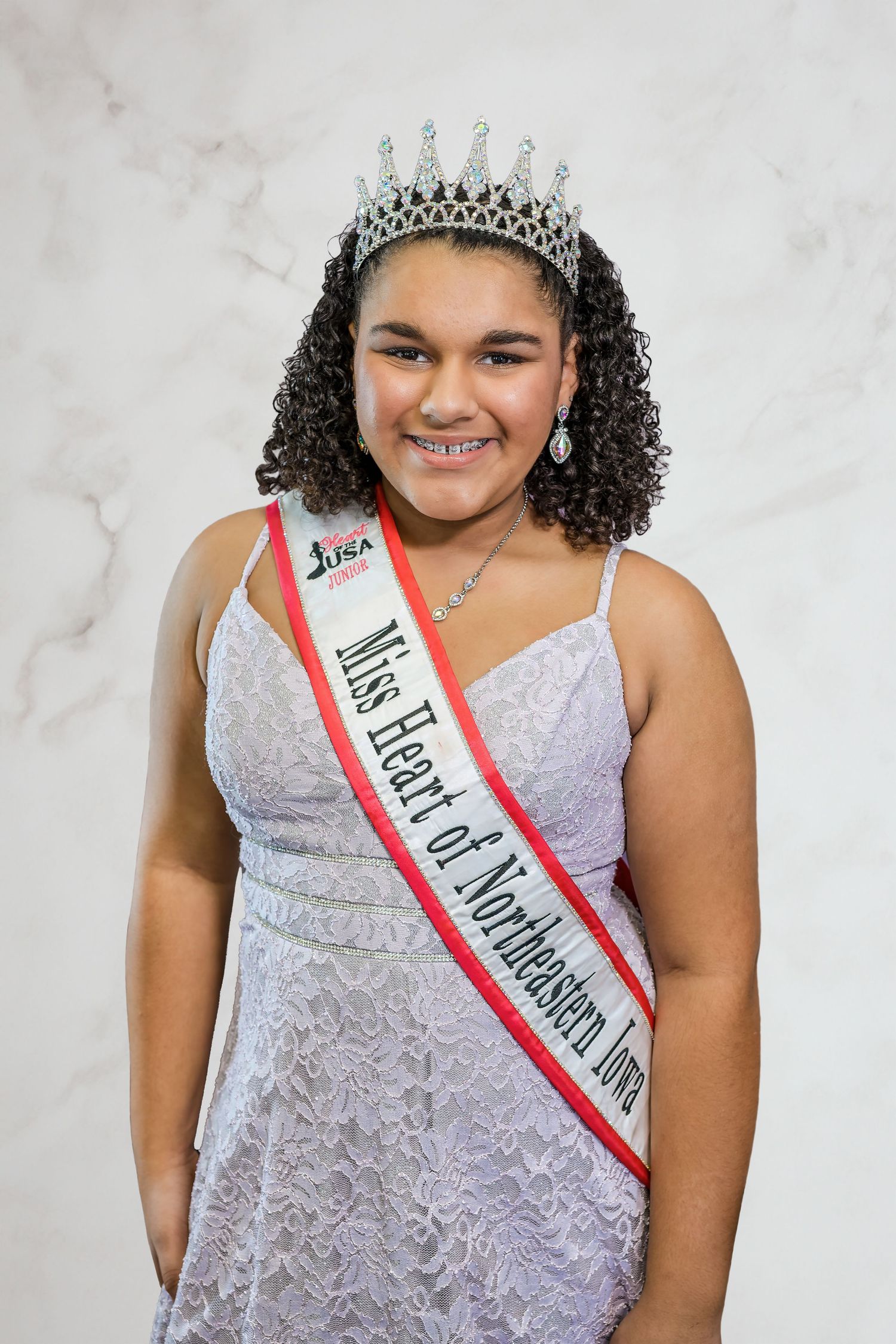 A beauty pageant contestant wearing a white lace dress, tiara, and red sash poses for a portrait against a gray background.