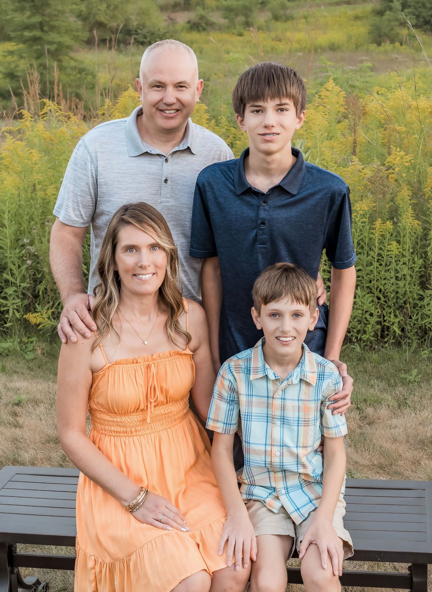 A family of four smiles for a portrait while sitting on a bench in front of yellow wildflowers on a summer day.