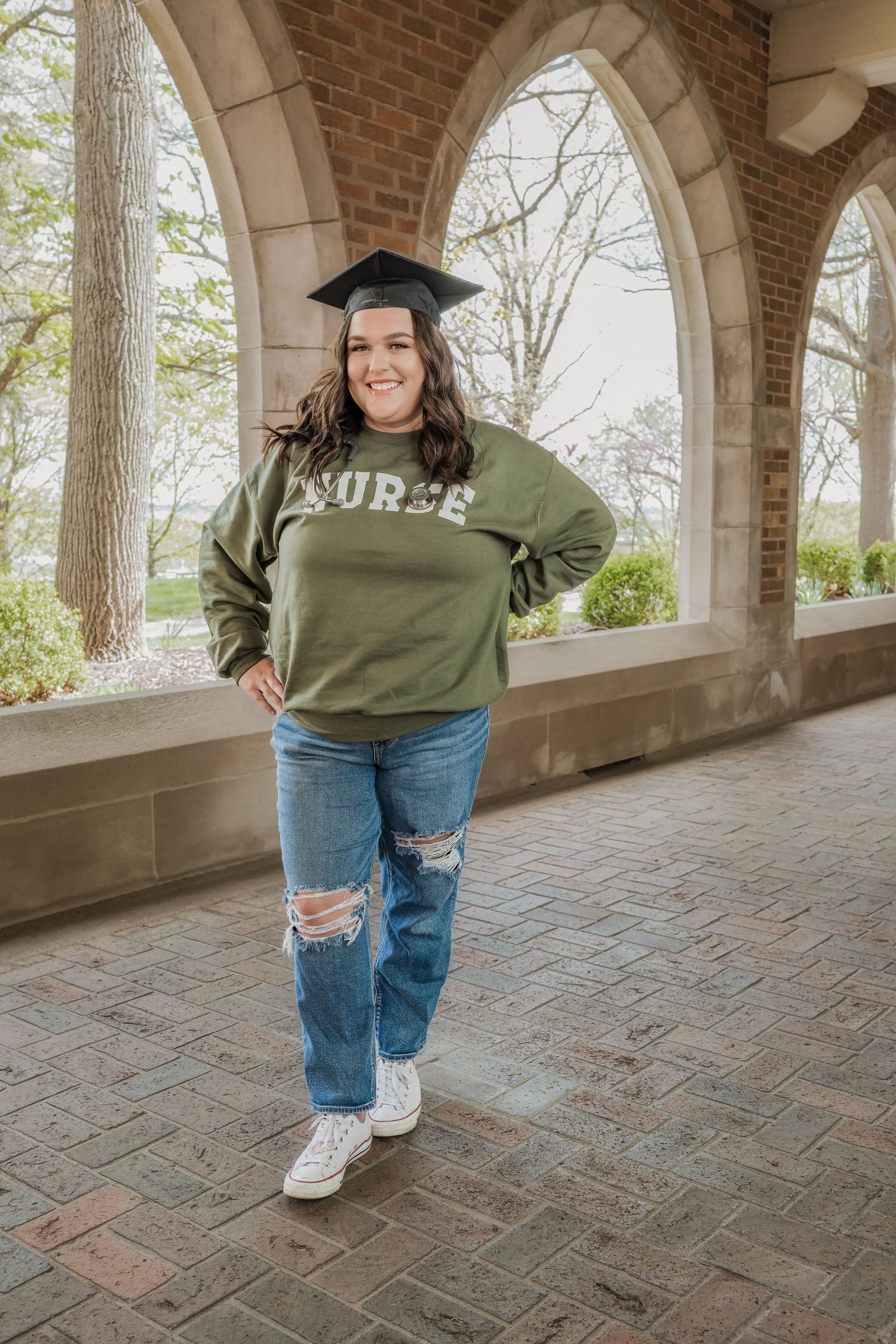 A graduate in a cap and olive green 'NURSE' sweatshirt poses under brick archways with distressed jeans.