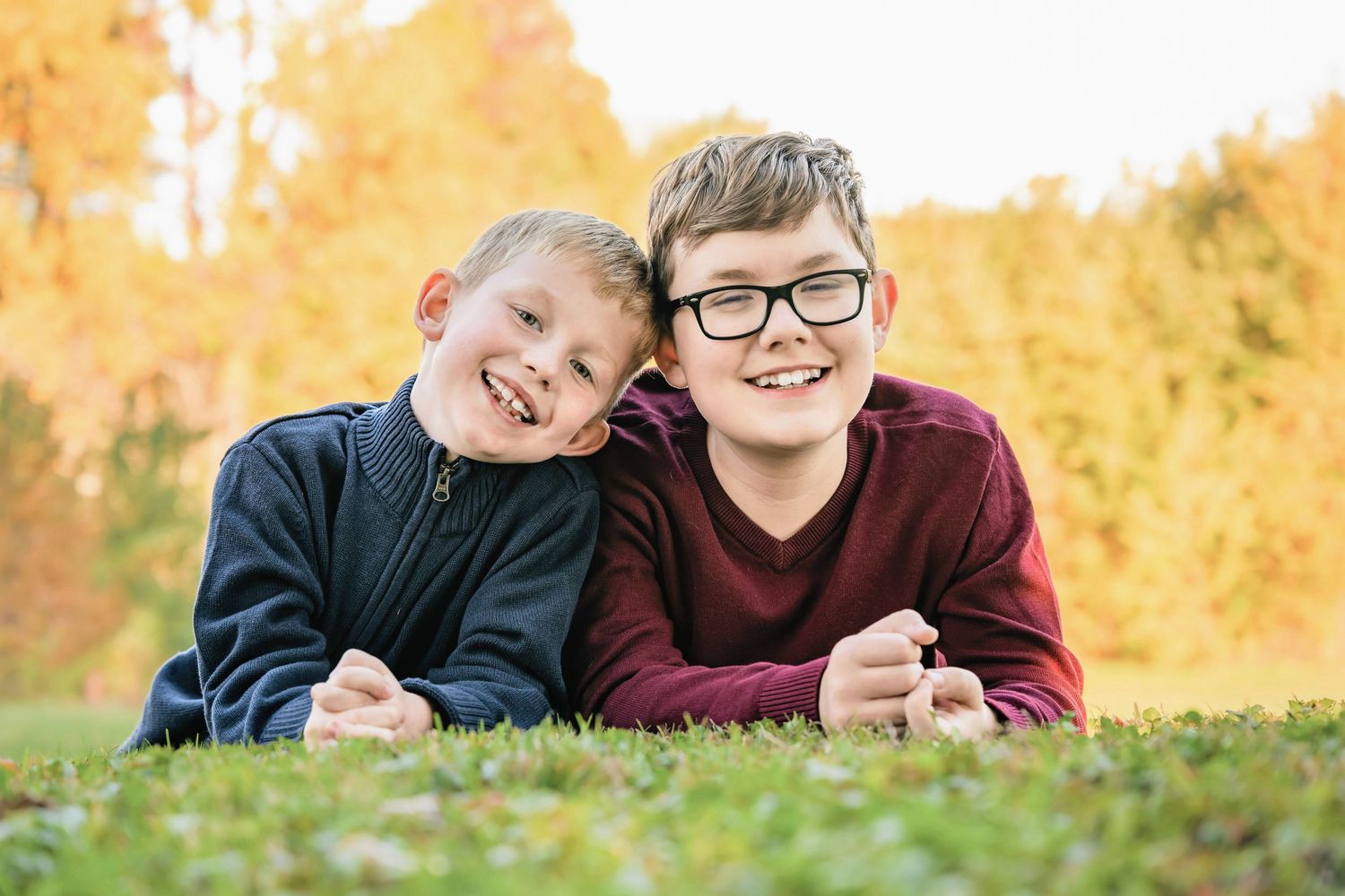 Two boys laying on grass during autumn with golden trees in background.