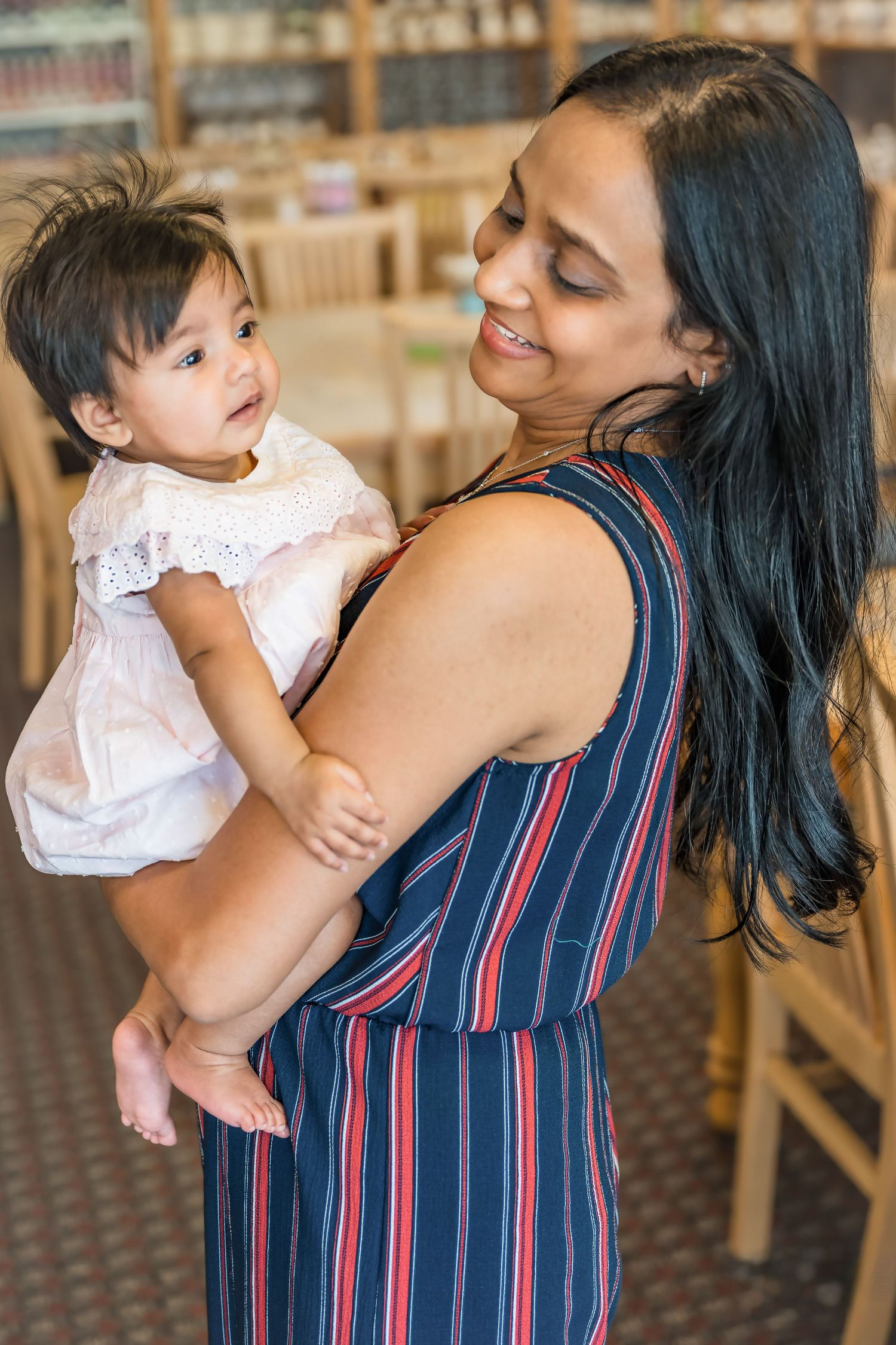Woman in striped dress holds smiling baby in white dress indoors.