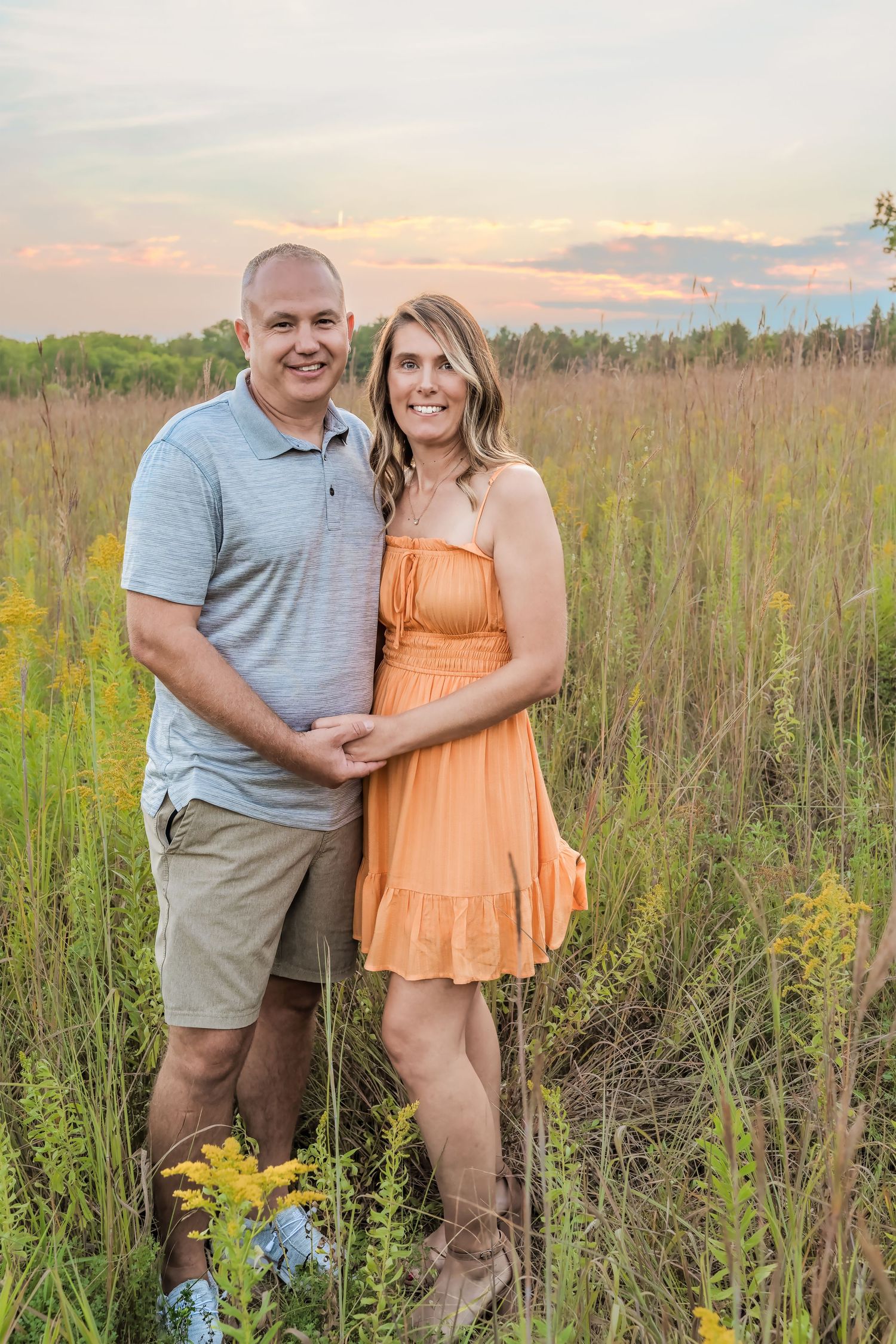 A couple stands together in a tall grass field during sunset, with wildflowers blooming around them.
