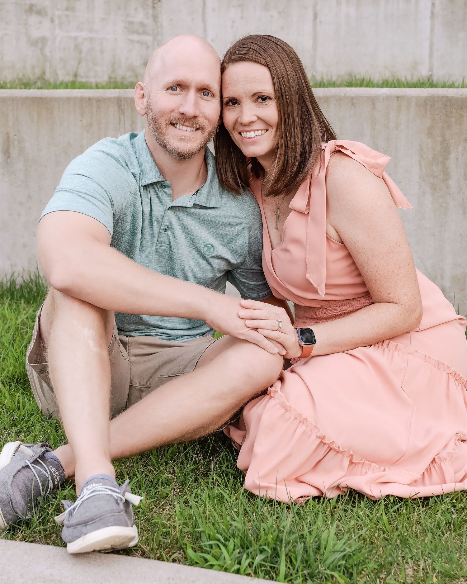 A couple sits together on grass wearing casual summer clothing in peach and mint tones against a concrete wall.