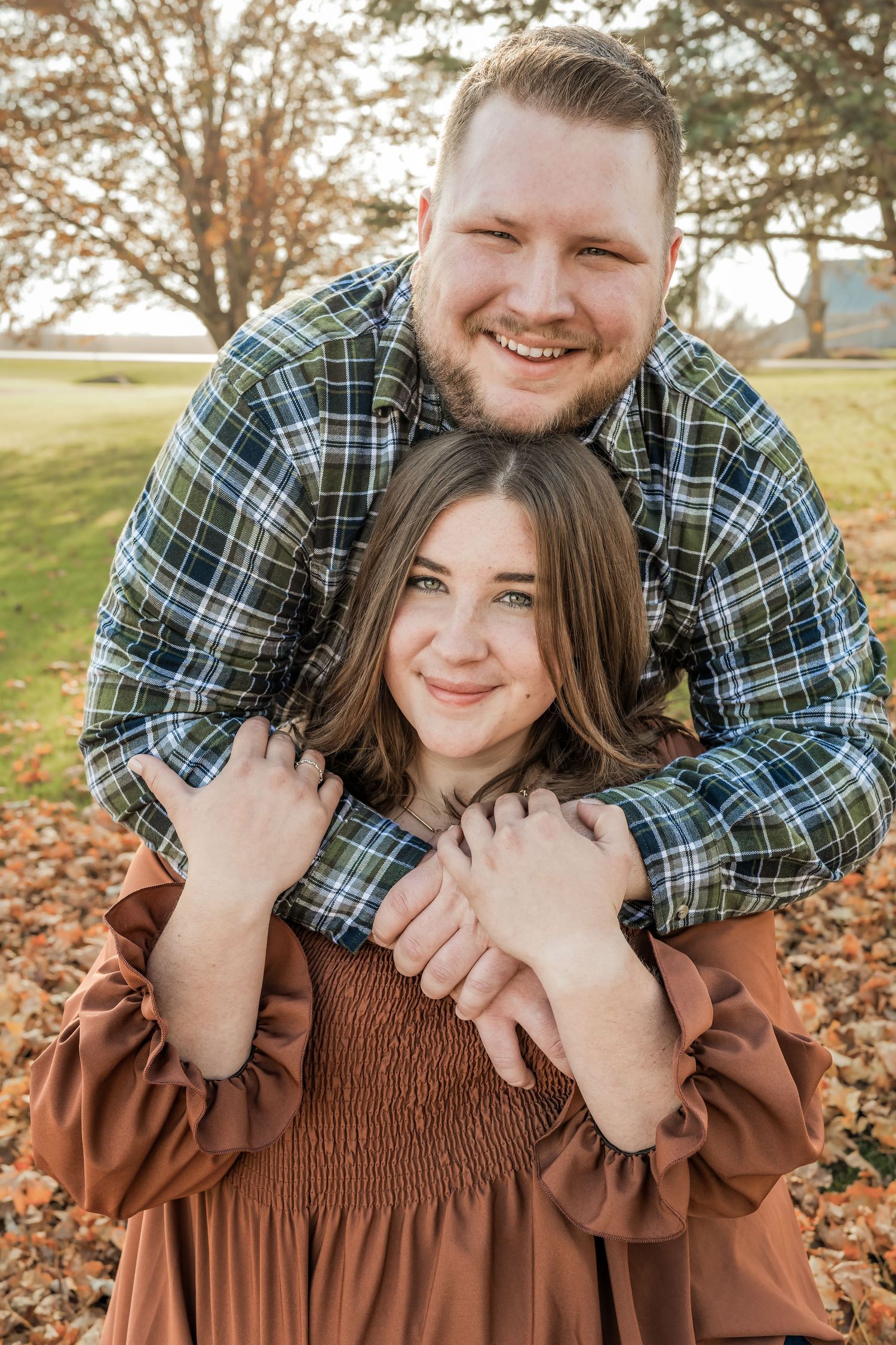 A couple embraces in an outdoor autumn setting with fallen leaves and sunlit trees in the background.