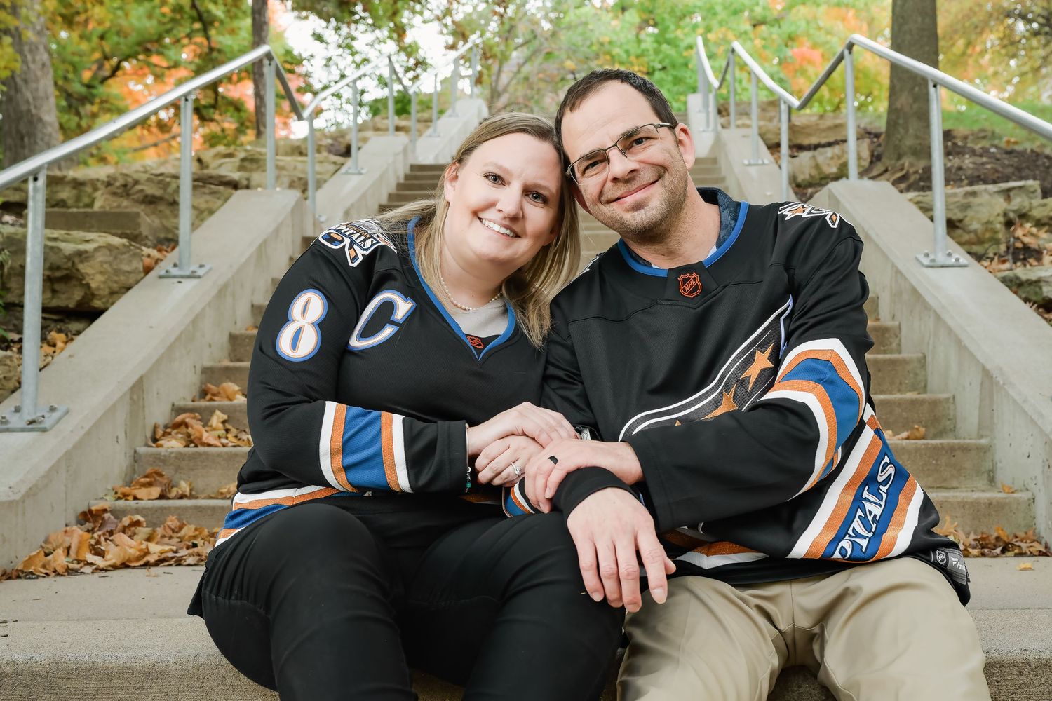 Two sports fans wearing matching team jerseys sit together on concrete steps during fall with autumn leaves visible.