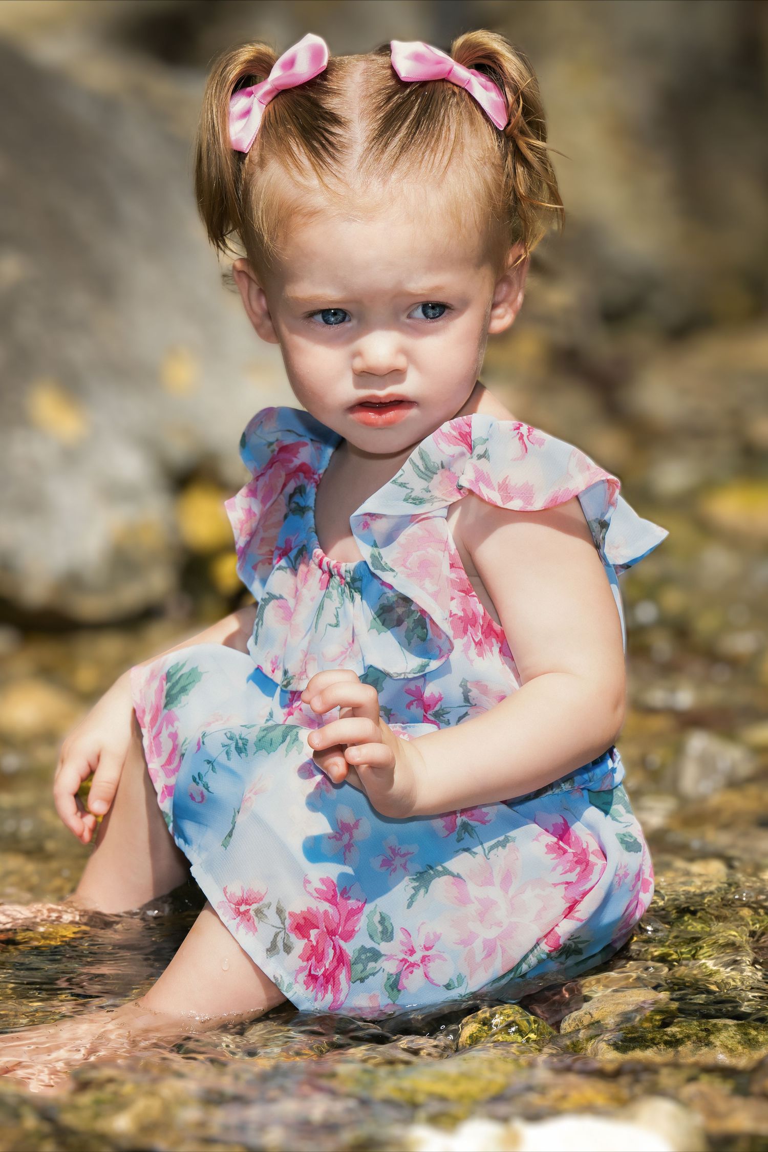 A toddler in a floral blue and pink dress with matching hair bows sits on a rocky surface outdoors.