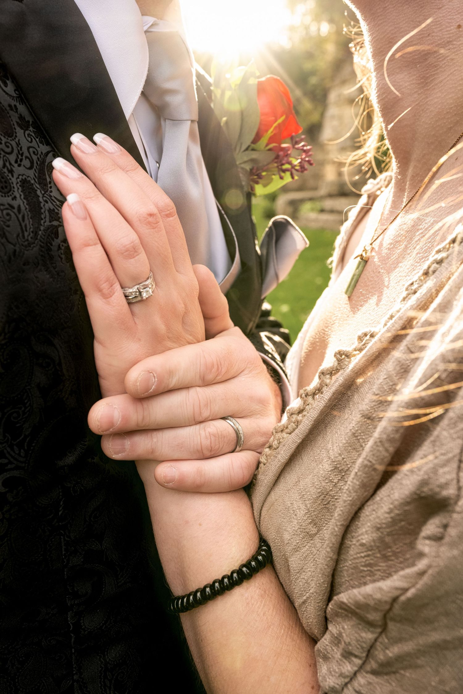 Close-up of joined hands showing wedding rings with a red rose and sunlight in the blurred background.