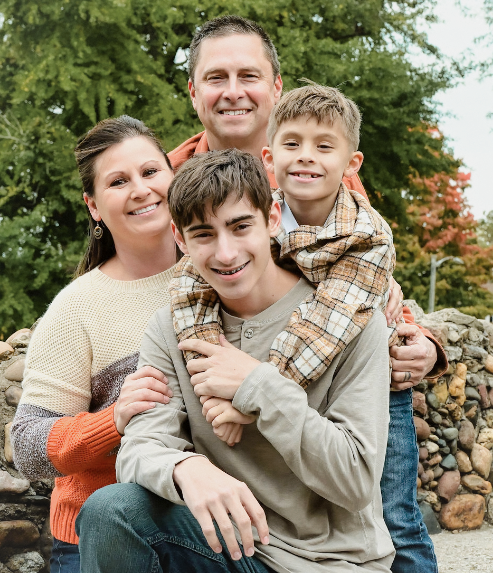 A family poses for a casual outdoor photo together against a backdrop of autumn trees and a stone wall.