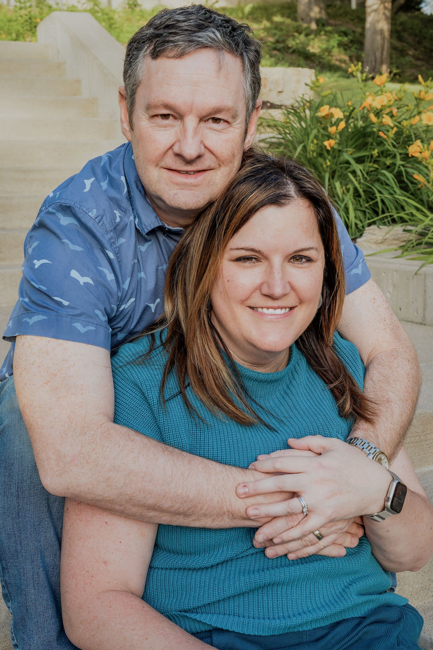 A couple sits together outdoors on steps surrounded by yellow daylilies, with one wearing a blue patterned shirt and one in teal.