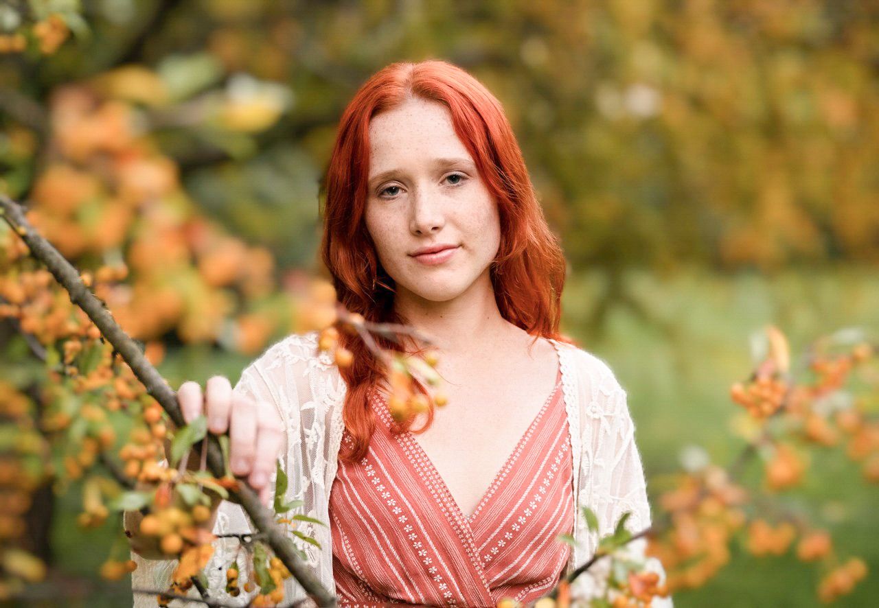 Person with vibrant red hair wearing a pink dress poses outdoors near autumn foliage with warm natural lighting.