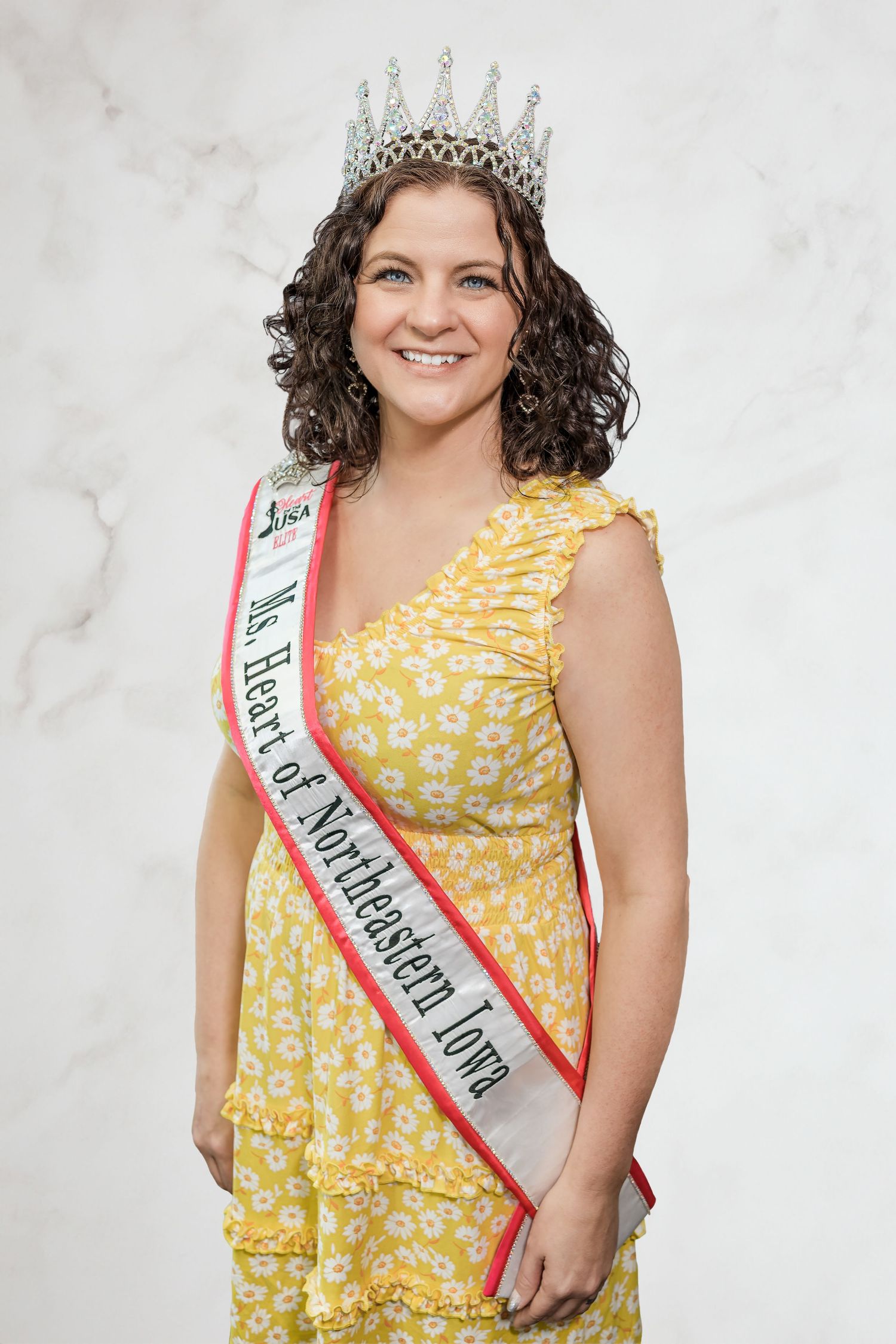 A pageant winner wearing a sparkling crown and yellow floral dress with a red and white sash.