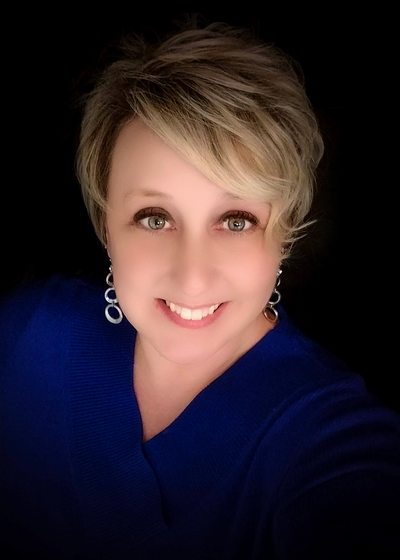 Professional headshot of a woman in a blue top against dark background with dangling earrings.