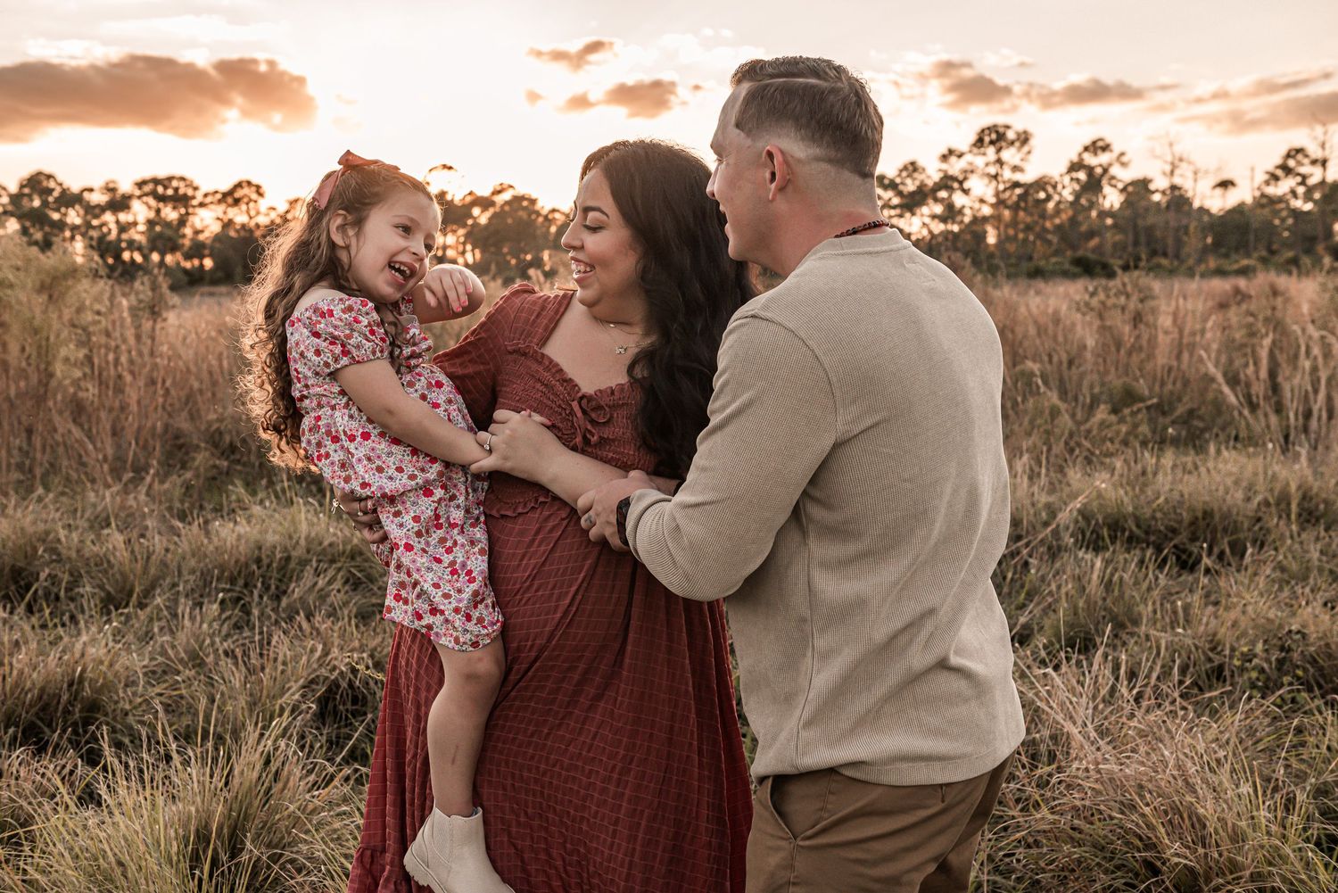 A family shares a tender moment together in a golden field at sunset with warm orange sky in the background.