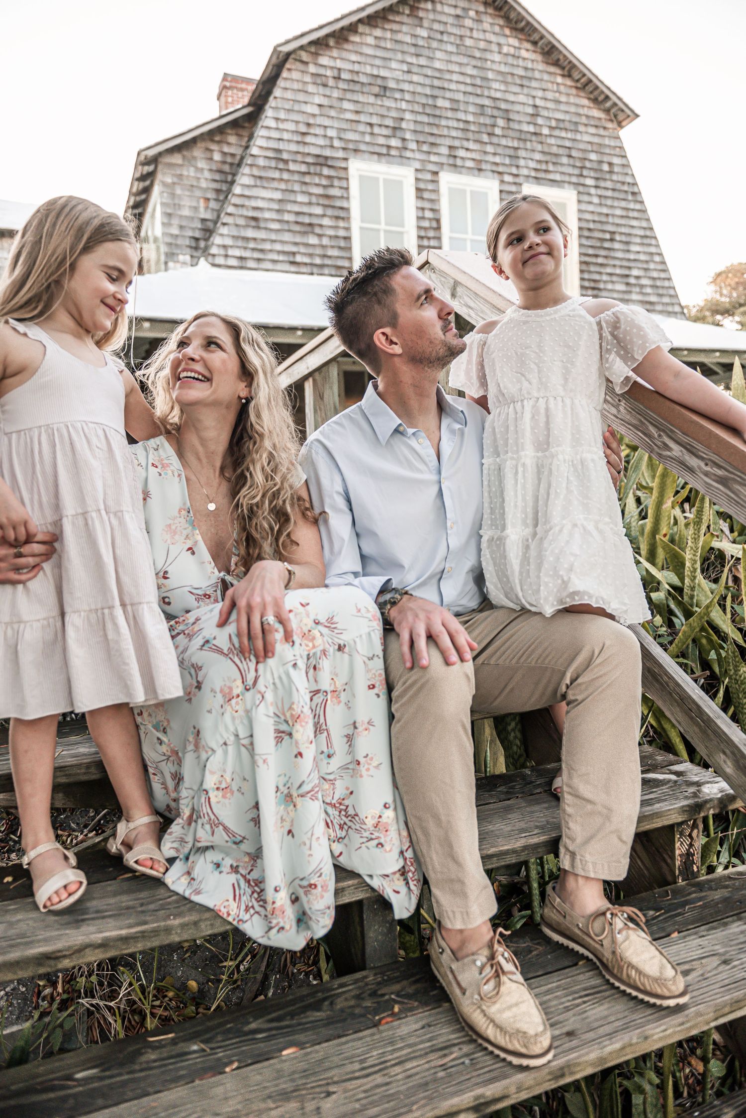 A family sits together on wooden steps outside a shingle-sided house, wearing coordinated casual summer outfits.