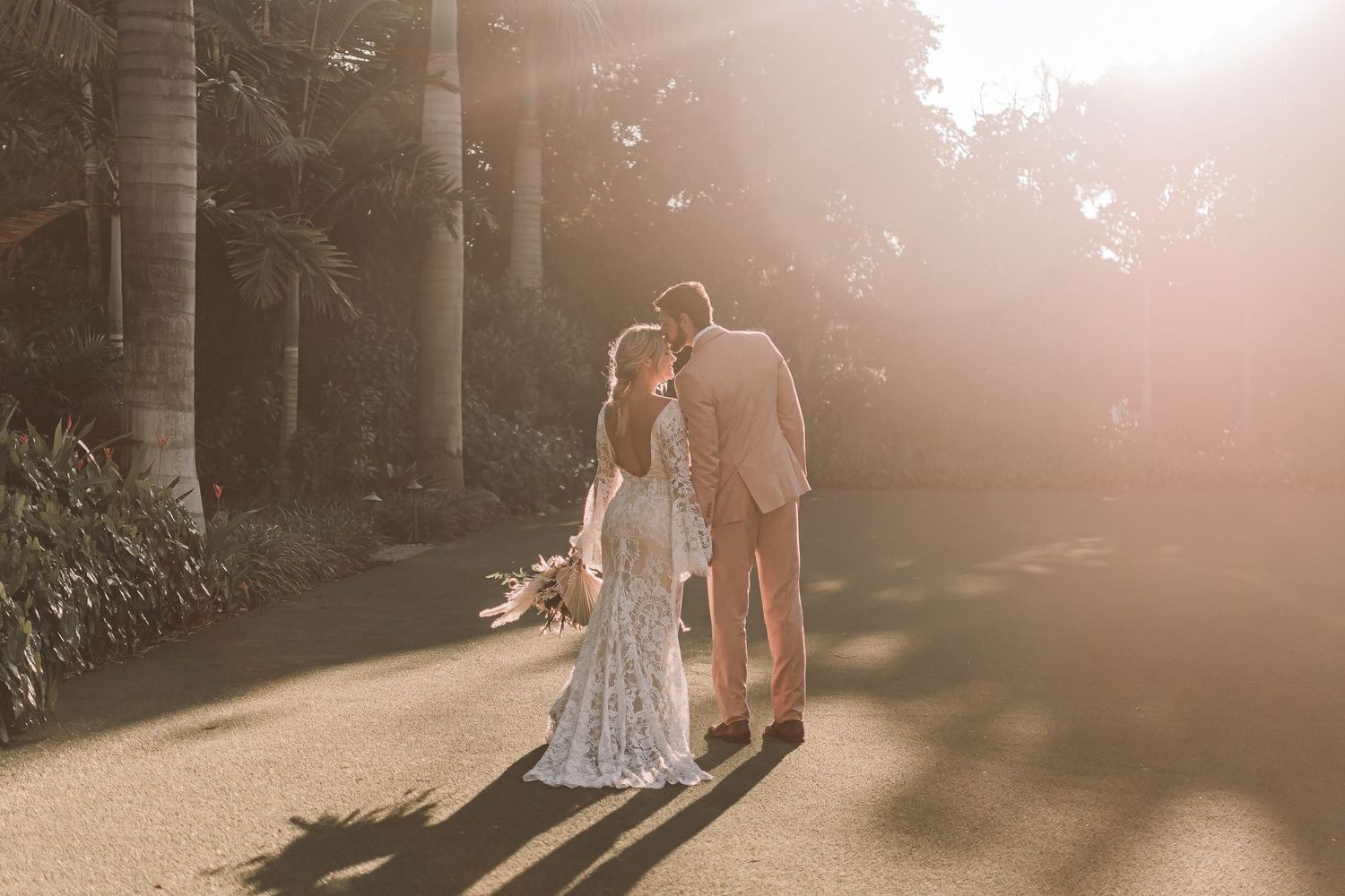 Couple walking hand in hand during sunset with long shadows cast on palm-lined path.