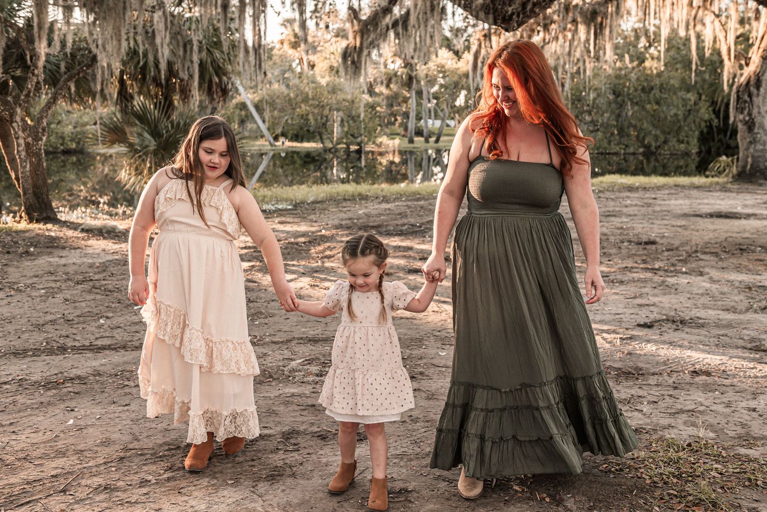 Three people in coordinating maxi dresses walk hand in hand through a mossy wooded area at sunset.