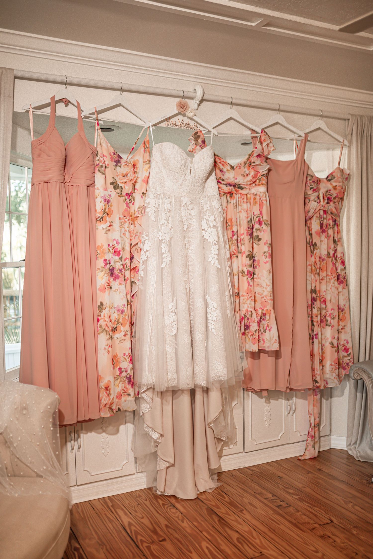 Wedding dress and pink floral bridesmaid dresses hanging on hangers against a white wardrobe in natural light.