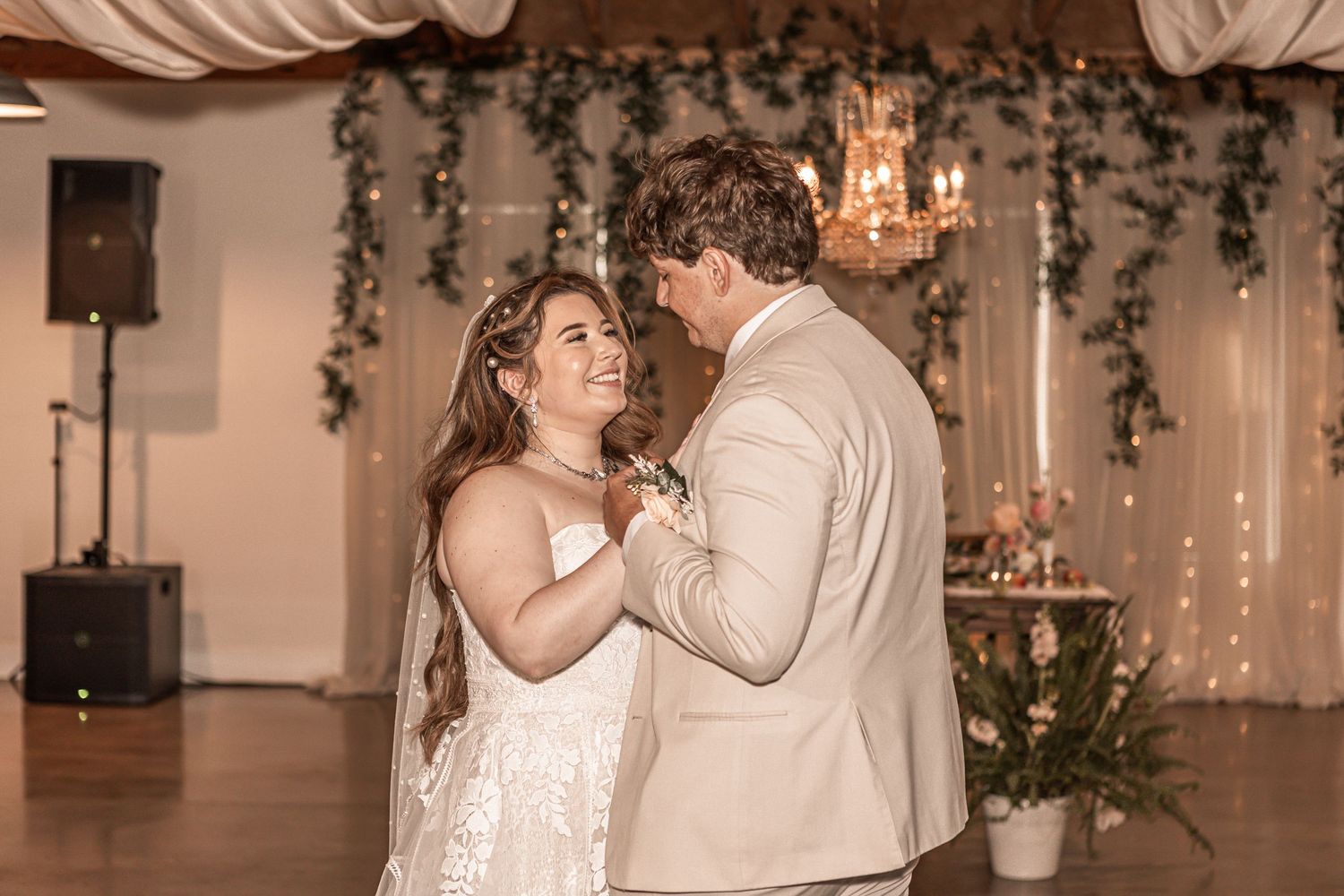 A couple shares their first dance at a wedding reception decorated with hanging greenery and chandeliers.